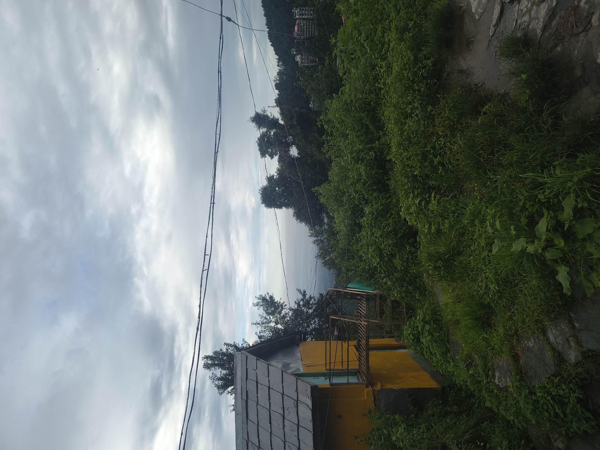 An overcast day in Haini, India, is depicted in this outdoor scene. In the foreground, a yellow building with a grey slate roof is partially visible on the left. A metal staircase with railings leads up to an entrance. Dense green foliage covers the ground around the building, with some areas appearing damp, possibly from recent rain. In the background, a landscape unfolds with lush green trees and a distant view of a hazy horizon, possibly a body of water. Several buildings, including a multi-story structure, are visible on a hillside in the mid-ground, suggesting a settlement or town. Overhead, power lines stretch across the frame, and a few distant birds are visible in the sky.