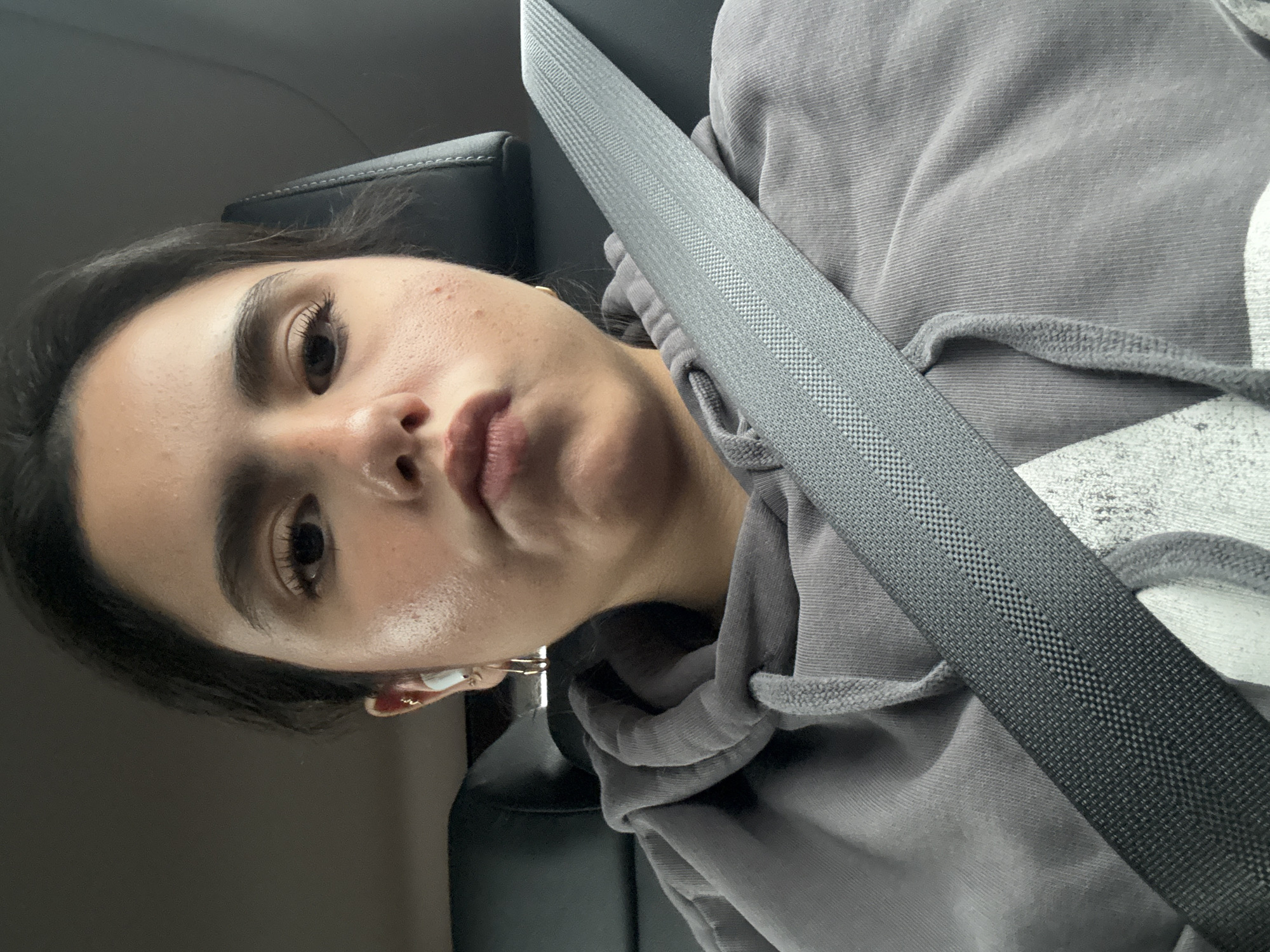 A close-up, slightly rotated selfie captures a young woman seated in the passenger seat of a car, looking directly at the camera. She has a somewhat pouting or serious expression. Her skin shows a natural texture with visible pores and a slight sheen, and a few minor blemishes are visible on her forehead and cheek. Her dark hair is pulled back from her face, highlighting her defined eyebrows and long eyelashes.
She is wearing a grey hooded sweatshirt, with the textured fabric visible around her. A dark grey car seatbelt diagonally crosses her body from her left shoulder downwards, its interwoven pattern clearly discernible. In her left ear, she wears a white wireless earbud and a small gold hoop earring. The interior of the car, including the headrest of the seat, is visible in the background, appearing dimly lit. The image suggests a candid moment, possibly taken while traveling or waiting in a car in Austin, United States of America. No text is visible in the image.