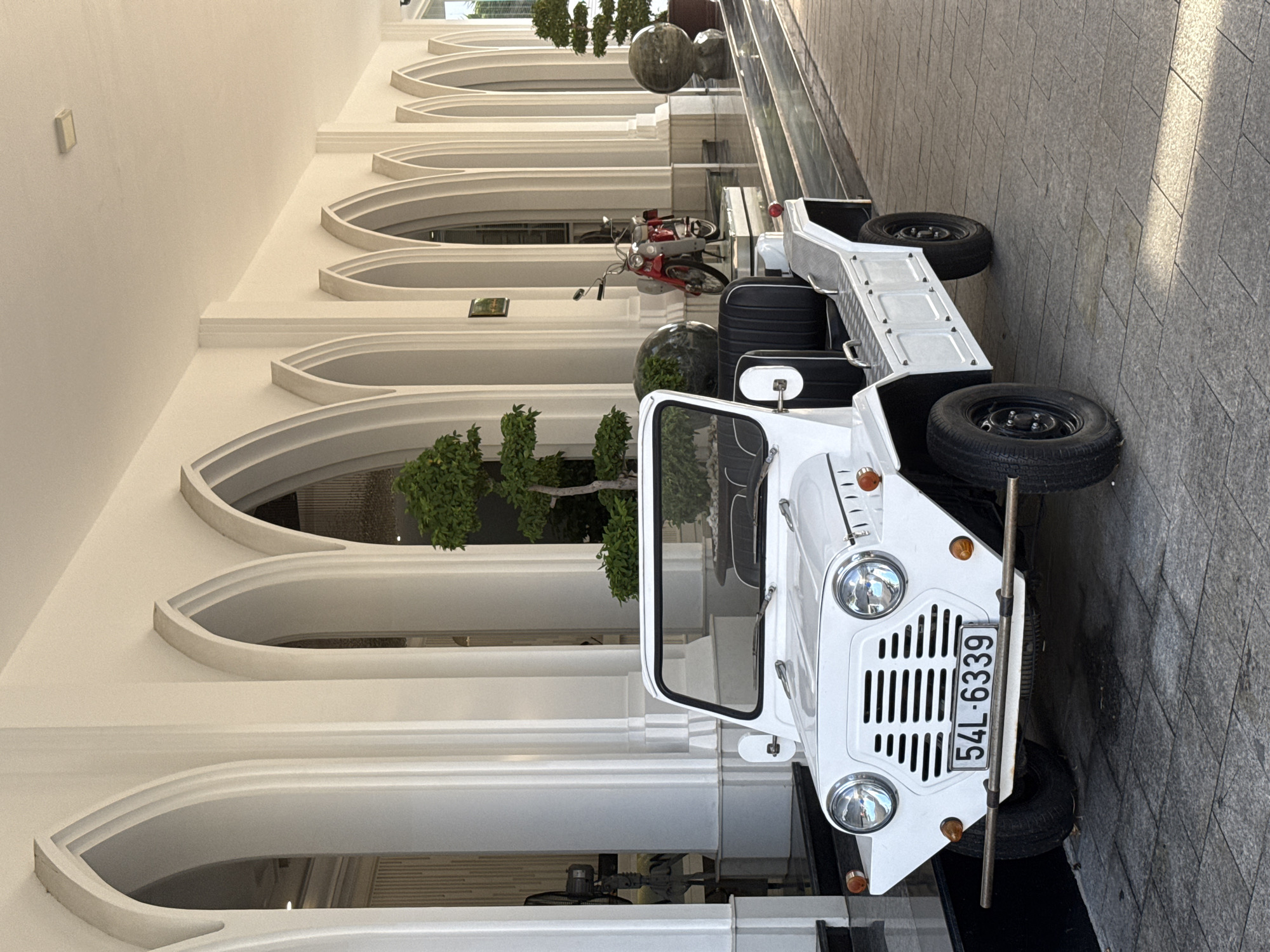 The image captures a white, open-top vehicle, resembling a classic Mini Moke, parked on a paved walkway in front of a building with a series of arched openings. The vehicle has a license plate that reads "54L-6339". In the background, beyond the vehicle, a red and silver moped is visible. The architecture of the building features white columns and arches, suggesting a hotel or a similar establishment. Lush green plants, including a bonsai-style tree, are placed strategically along the walkway, adding a touch of greenery to the scene. The ground is paved with rectangular stone tiles. Sunlight is casting shadows, indicating it is daytime. The overall setting appears to be an outdoor area of a resort or a hotel in Tay Nha Trang Ward, Vietnam.