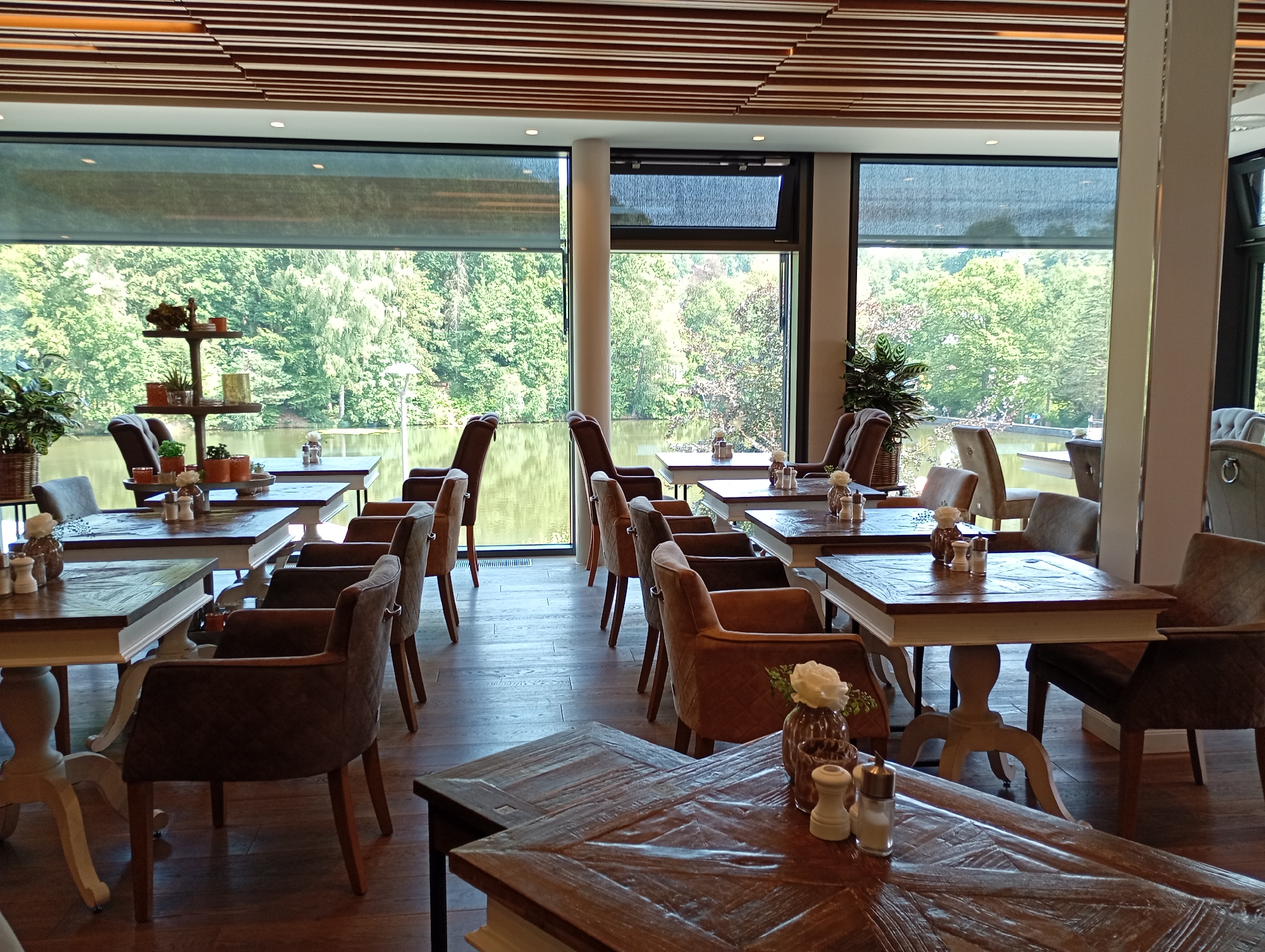 The image depicts the interior of a restaurant with a view of a lake and trees through large windows. The restaurant features wooden tables and upholstered chairs. In the foreground, two wooden tables are set with salt and pepper shakers, and a vase with a white flower. The tables are made of dark wood with a carved surface. Several chairs surround the tables, upholstered in brown and gray fabric. The windows offer a scenic view of a body of water and lush green trees. The ceiling is constructed with wooden slats. In the background, more tables and chairs are visible, as well as a decorative shelf with potted plants and figurines. The scene appears to be a dining area within the restaurant, likely the "Forellenhof am Sösetal," a restaurant located in Bad Sachsa, Germany, known for its scenic setting by a lake. No people are clearly visible in the image.
