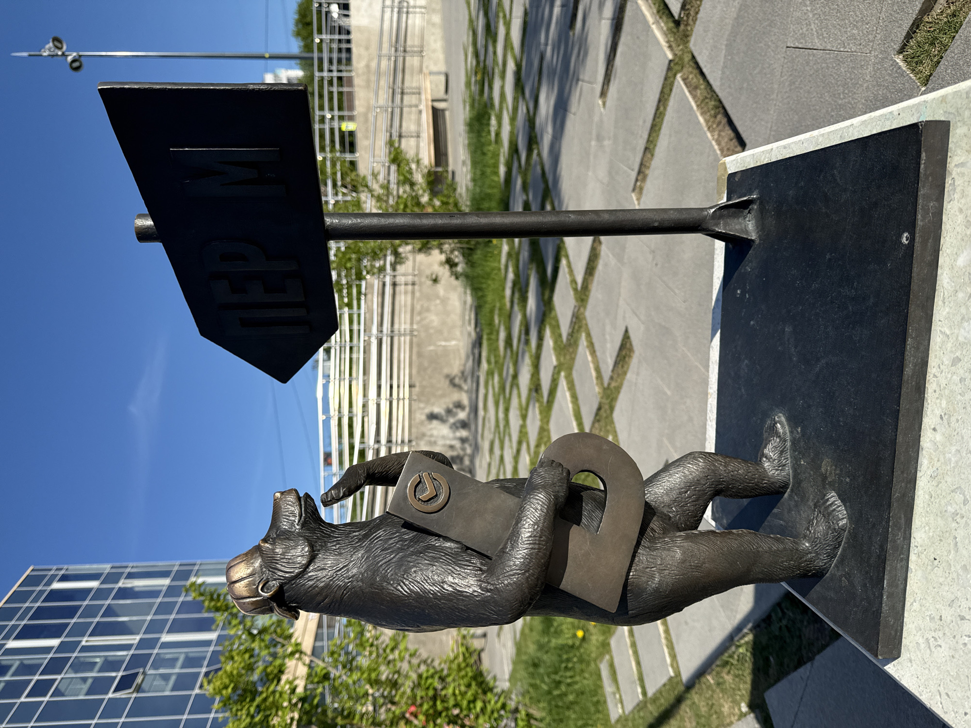 The image shows the "Permyak Salty Ears" sculpture in Perm, Russia. The sculpture consists of a bronze bear on a black base holding a letter "П" and a frame with a "У" inside, with a sign above reading "ПЕРМЬ" (Perm). The sculpture is located outdoors in a public square with gray and white paving stones. A building and some scaffolding are visible in the background under a blue sky.