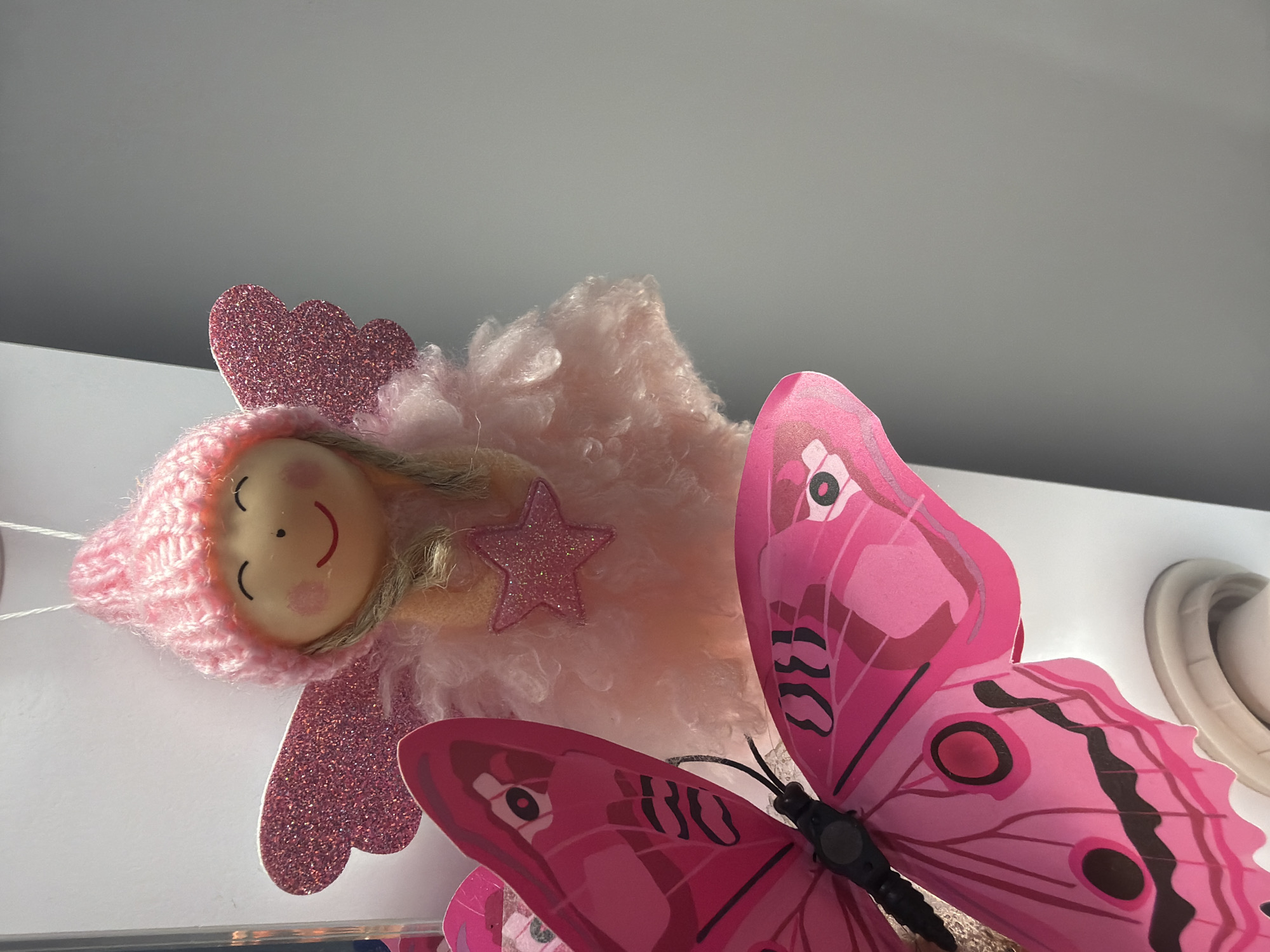 A close-up, low-angle shot shows a pink knitted doll resembling an angel with glittery wings and a small star ornament. In the foreground, a large pink butterfly decoration is prominently displayed, with its wings partially obscuring the doll. The background is a neutral gray wall. The setting is domestic, likely an indoor decorative display, with no specific identifiable landmarks to place it within Kyiv, Ukraine.
