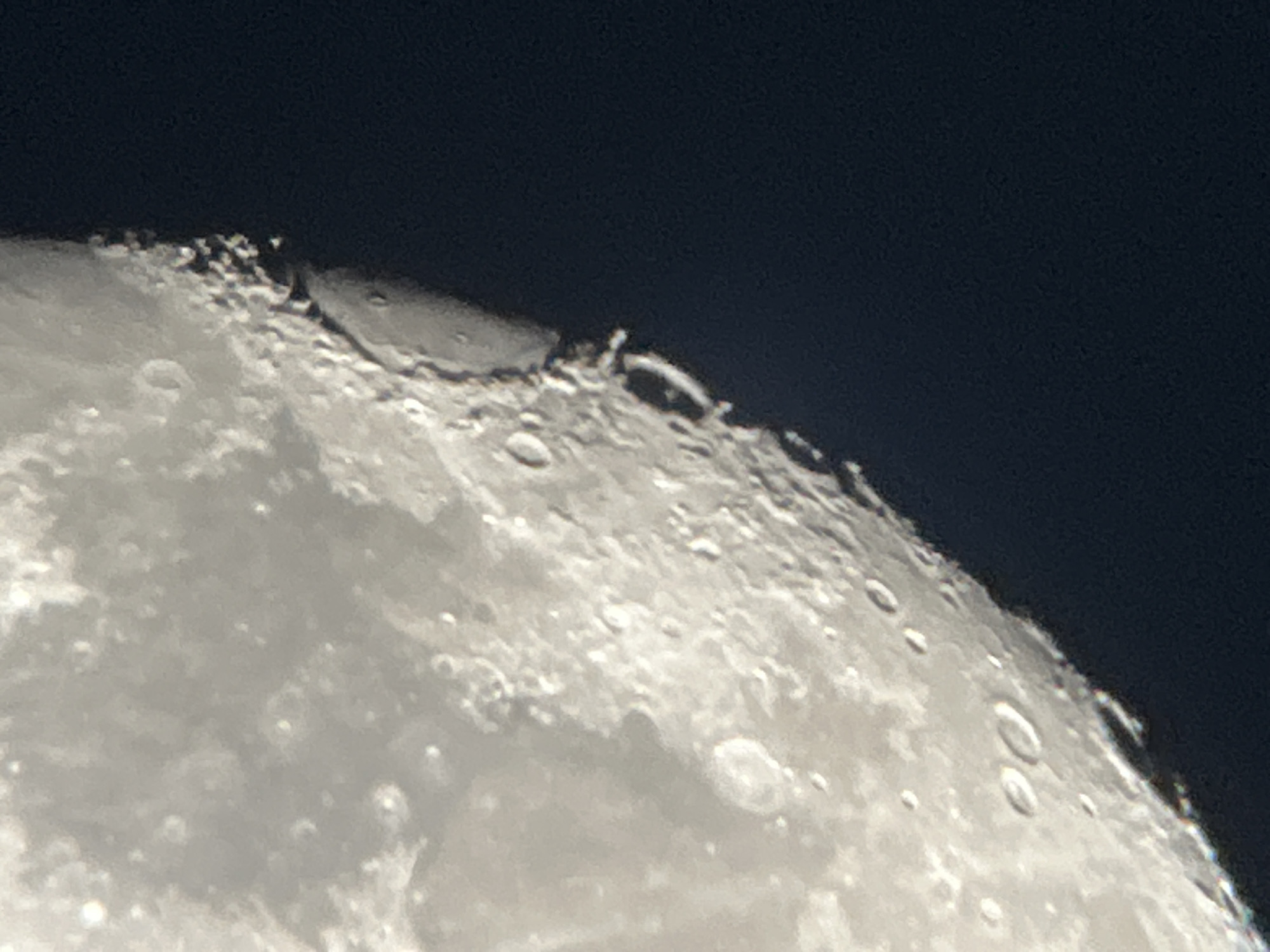 The image shows a close-up, detailed view of the Moon's surface. The lunar terrain is rugged and uneven, characterized by numerous craters of varying sizes, some appearing as bright circular indentations and others as darker shadows. The edge of the Moon is sharply defined against a dark, black background, which represents outer space. The surface is illuminated, highlighting the mountainous texture and the rims of the craters. There is a subtle spectrum of light and shadow, suggesting the angle of illumination. The image does not contain any elements related to the city of Lyubertsy, Russia, or any human presence.