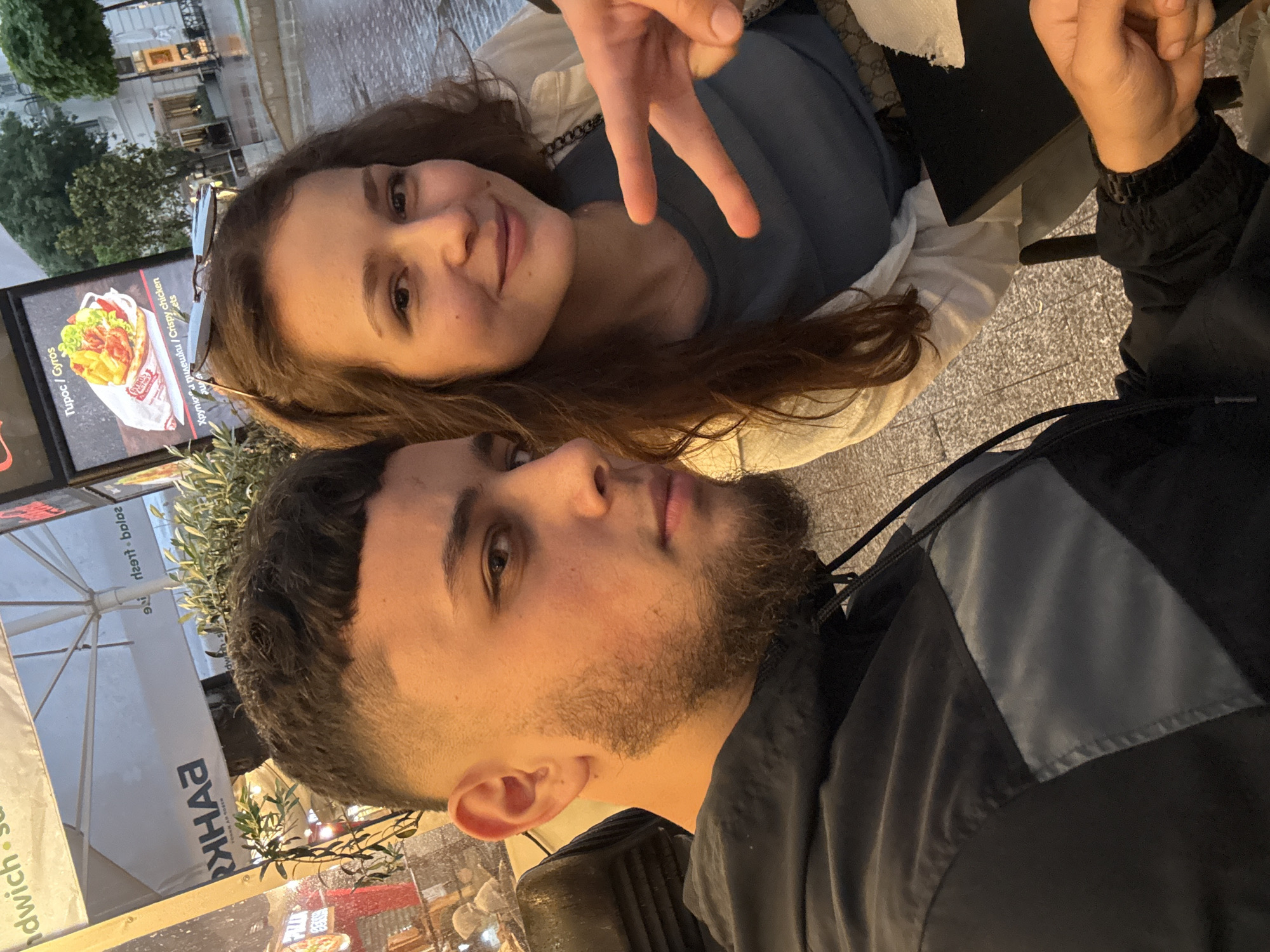 The image captures two young individuals, a man and a woman, posing for a photo in an outdoor urban setting in Varna, Bulgaria. The woman is positioned in the background, smiling cheerfully at the camera. She has long brown hair, wears a blue top and a light-colored jacket, and her right hand is making a peace sign. Sunglasses are perched on her head, and a strap with a possible Gucci pattern is visible near her shoulder.
In the foreground, the man looks directly at the camera with a calm, engaged expression. He has short dark hair, a neatly trimmed beard, and is dressed in a dark jacket with grey paneling.
The scene suggests they are at an outdoor cafe or restaurant. A prominent menu board displays Greek text, including "ΤυροC / Gyros" (Cheese / Gyros) and "Χοιρινo / Κεικενια / Crispy chicken" (Pork / Chicken / Crispy chicken), along with a price of "4.5". An umbrella in the background features the name "ВАНКO" (Vanko) and partially visible English words like "salad" and "sandwich." Green plants are also visible, contributing to the outdoor atmosphere. The ambient lighting suggests it is likely late afternoon or early evening. Both subjects appear relaxed and content.