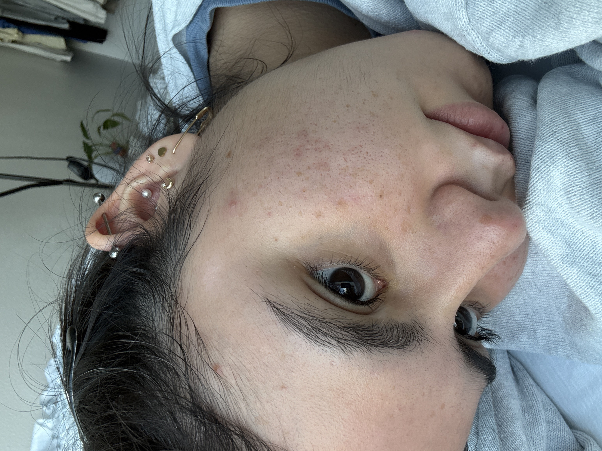 A close-up, side-profile image captures a person, likely a young adult, lying down with their head slightly tilted and gaze directed towards the viewer. The image is rotated 90 degrees counter-clockwise. The subject's dark hair frames their face, with some strands casually falling around their right ear (on the left of the frame).
The ear is adorned with several piercings, including a gold-colored bar in the helix, a daith piercing featuring a small gold hoop, a tragus piercing with a clear gem, a conch piercing with a pearl or silver stud, and two small gold studs in the lobe. The subject's skin is light-toned with visible freckles across the cheeks and forehead, along with some subtle redness and minor textural variations.
Their dark-irised right eye is open, making direct contact with the camera, complemented by long, dark eyelashes. A portion of the nose and naturally pink-hued lips are also visible. The person is partially covered by a soft, light grey fabric, possibly a blanket or garment, suggesting a comfortable, indoor setting. The background is softly blurred, revealing indistinct shapes that hint at an indoor environment, such as plants or furniture.
The lighting is soft and diffused, indicating natural daylight without harsh shadows, contributing to a calm and candid atmosphere. The subject's expression appears neutral and relaxed. No text is visible in the image. The provided location context for this media is Austin, United States of America.