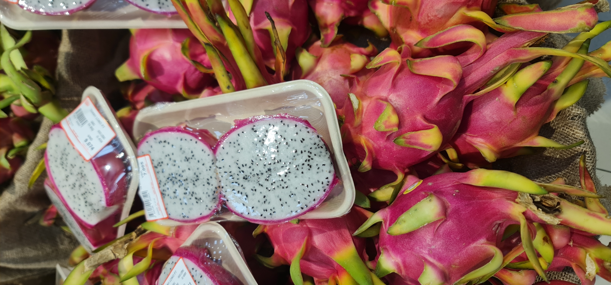 The image shows a display of dragon fruit, both whole and sliced, at a market in Đà Nẵng, Vietnam. Several whole, bright pink dragon fruits with green-yellow tipped scales are piled together. Two clear plastic trays hold dragon fruit halves, their white flesh speckled with tiny black seeds. Price stickers in Vietnamese currency (VND) are affixed to the trays. A burlap-like material serves as a base under the fruits. The lighting is bright, suggesting an indoor market setting.