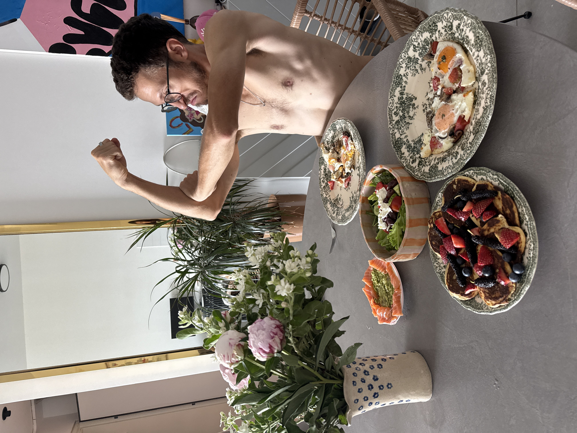 A shirtless, light-skinned man with dark hair and glasses stands beside a round gray table laden with food. He flexes his left arm. The food includes fried eggs, a salad, salmon with avocado, and pancakes with berries, all served on floral-patterned plates and dishes. A bouquet of flowers in a blue and white vase sits at the edge of the table. The background includes a rattan chair, a potted plant, and a mirror. The location is suggested to be an apartment in Ramat Gan, Israel, although there's no explicit visual indication of this.
