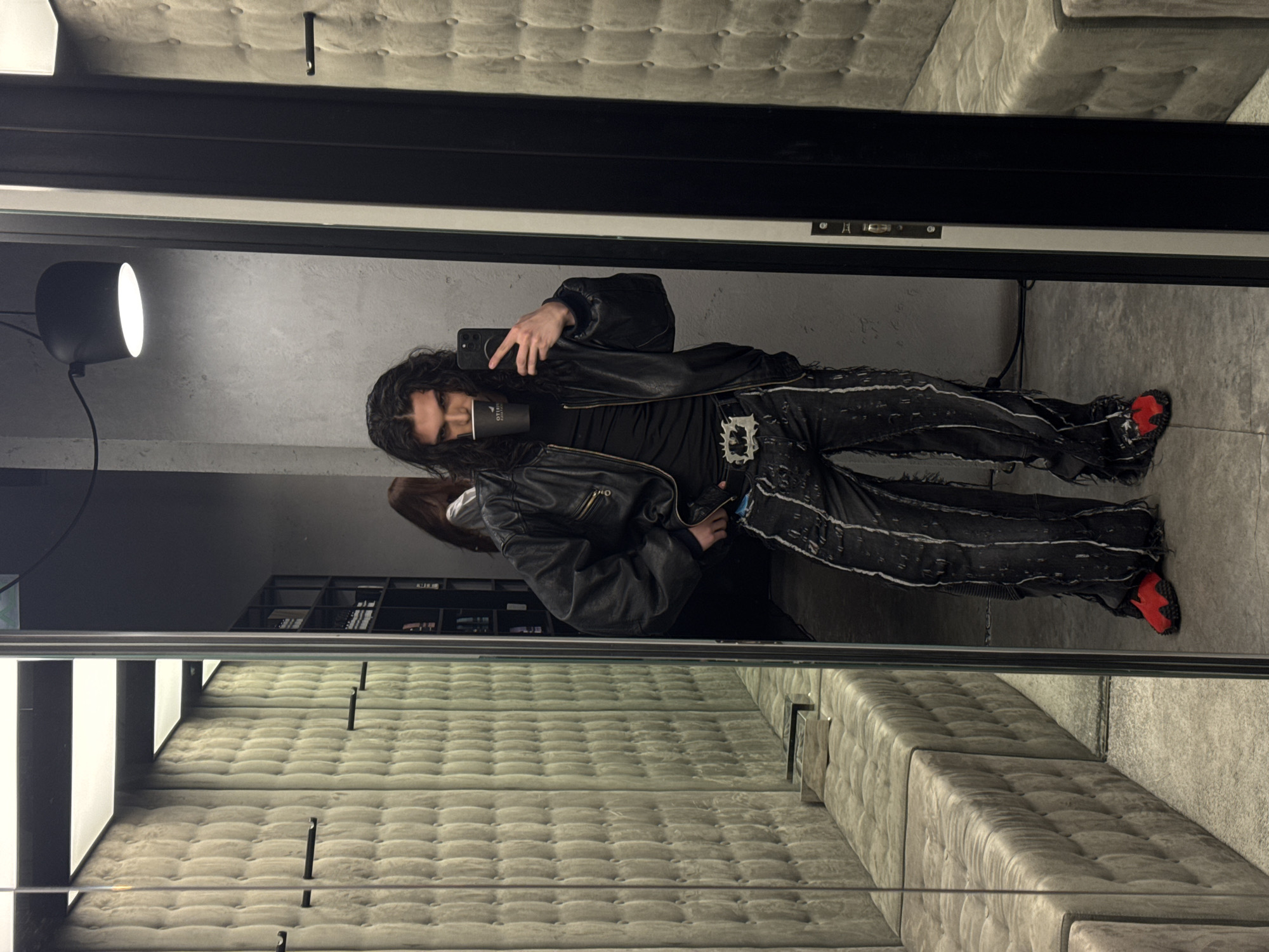 A long-haired individual, appearing male, is captured taking a mirror selfie inside what resembles a modern fitting room or retail lounge. The individual wears a black leather bomber jacket, a black t-shirt, dark distressed wide-leg jeans with white markings, a prominent silver belt buckle with a jagged design, and red textured slip-on shoes. They hold a black smartphone in their left hand, angled to capture their reflection, and a black cup with a white logo to their mouth with their right hand.
The immediate environment visible in the reflection features light beige or grey quilted upholstery on the wall to the left, with matching upholstered benches below it and black clothing hangers mounted on the wall. The floor is a light grey, possibly concrete. Beyond the mirror, the actual space consists of dark grey textured walls, a black modern pendant light fixture, and a dark shelving unit displaying items. A second individual with long brown hair and a white shirt is partially visible in the reflection, standing further back and to the left of the primary subject, facing away. The exact location in Sofia, Bulgaria, cannot be determined from the visual information provided.