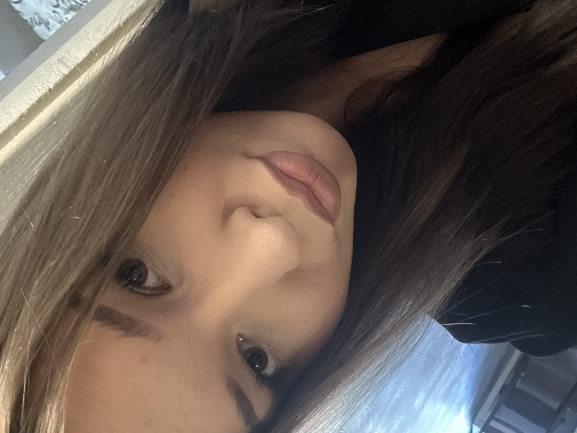 The image is a close-up, selfie-style portrait of a young woman. She has long, brown hair that is parted and falls around her face. Her right eye, with dark brown iris and visible eyelashes, is in focus, along with her nose and lips. Her lips are painted with a matte, rosy-brown lipstick. The background is out of focus, showing hints of a textured wall and possibly a window with a blue tint. The lighting appears to be artificial and casts a soft glow on her face. There is no indication of the city of Inskoy, Russia, within the image or its context.