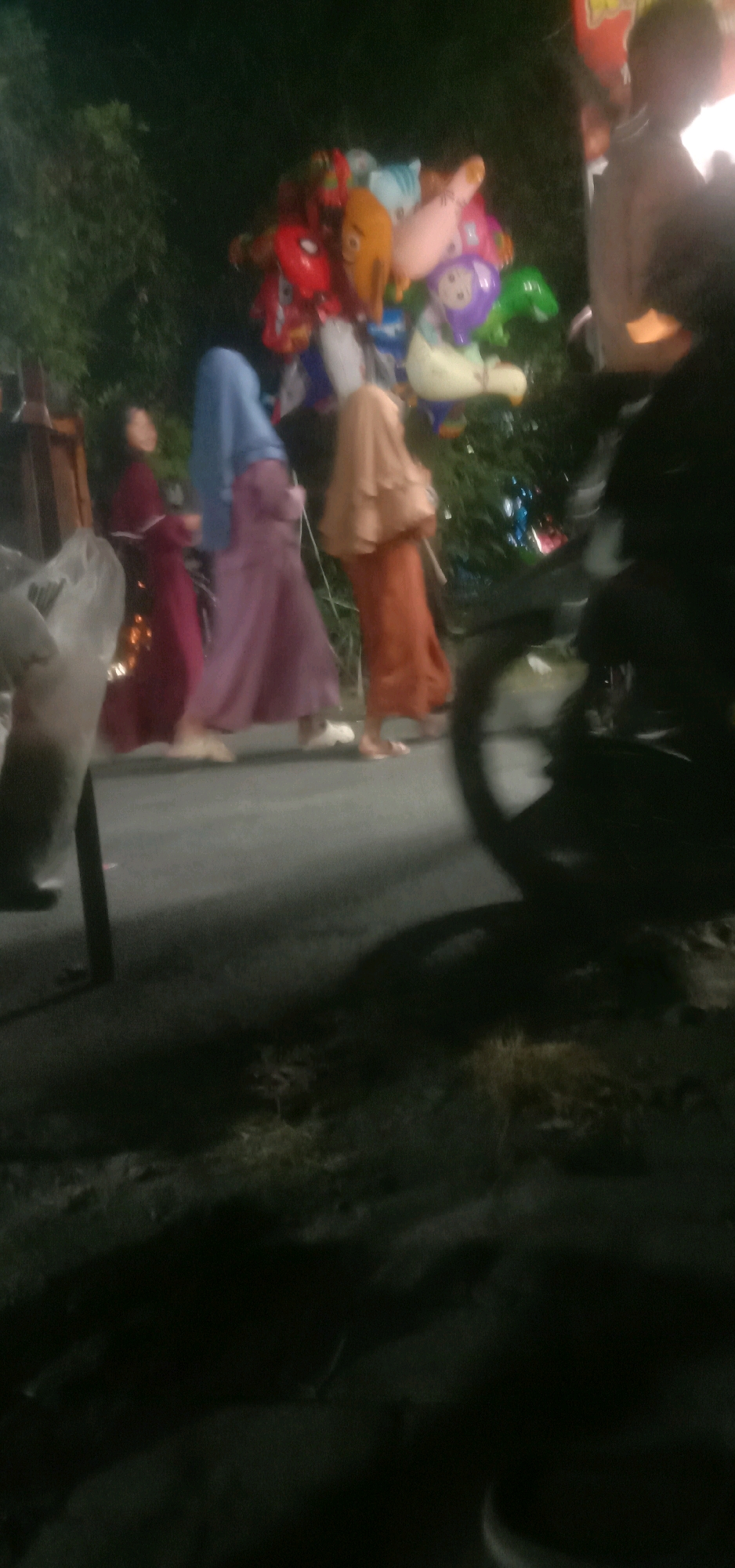 A group of people are walking on a street at night in Banjar Dowo, Indonesia. The scene is dark, with some artificial lighting present, casting shadows. In the foreground, a motorcycle is visible on the right side of the image, partially obscuring the view of the street. A person is seated on the motorcycle, and their silhouette is visible.
To the left of the motorcycle, a cluster of colorful balloons, some shaped like cartoon characters, are being held. Three individuals are walking past the balloons. The person closest to the balloons is wearing a pink hijab and an orange dress. To their left, another person is wearing a blue hijab and a purple dress. The person furthest to the left is wearing a dark maroon dress and has dark hair. They appear to be walking away from the balloons.
The ground is paved, and there is vegetation visible in the background, suggesting an outdoor setting. The overall atmosphere is that of a lively street scene at night, possibly during a festival or market.