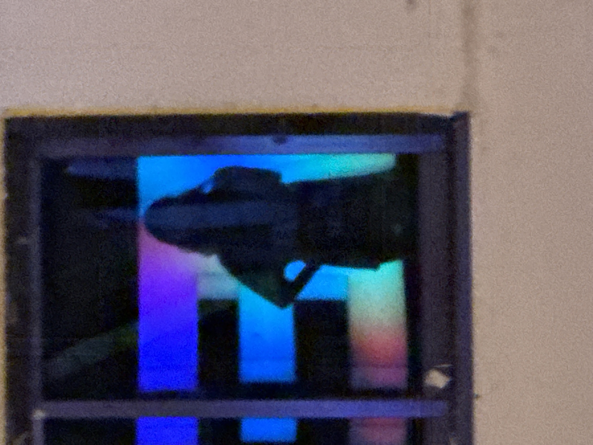 A silhouette of a person, appearing to be a woman, is visible through a window. The person is standing with their right hand on their hip. The window has a grid pattern and is illuminated from behind with vibrant, abstract colors, primarily blues and purples, with hints of pink and green. The lighting creates a stained-glass effect. The scene is set within a building with a light-colored exterior wall visible to the right of the window. The image appears to be taken at night. The location, ตำบลเกาะพะงัน, Thailand, suggests this could be a scene from a bar, club, or residential window on the island.