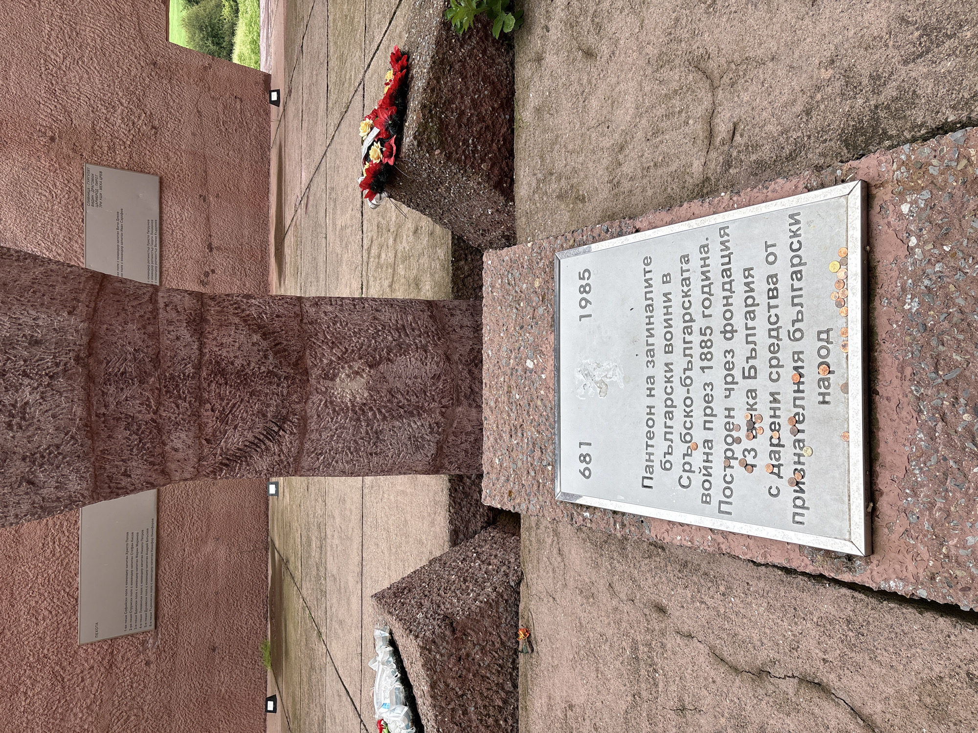 The image depicts an outdoor monument, likely in Gurgulyat, Bulgaria. In the foreground, a metallic plaque is mounted on a rough, reddish-pink stone base. The plaque is inscribed with text in Bulgarian, with the numbers "681" and "1985" prominently displayed at the top. Various coins are scattered on the plaque and the surrounding stone. Behind the plaque, the monument features large, rough-hewn, reddish-pink stone structures, reminiscent of columns or abstract sculptures. To the left and right of the central stone structures, there are angled stone blocks serving as bases. A wreath of red and yellow flowers is placed on the stone block to the right. In the background, parts of a textured reddish-pink wall are visible, with some inscriptions or plaques mounted on it. The ground appears to be paved with stone slabs. The overall impression is one of a memorial site.