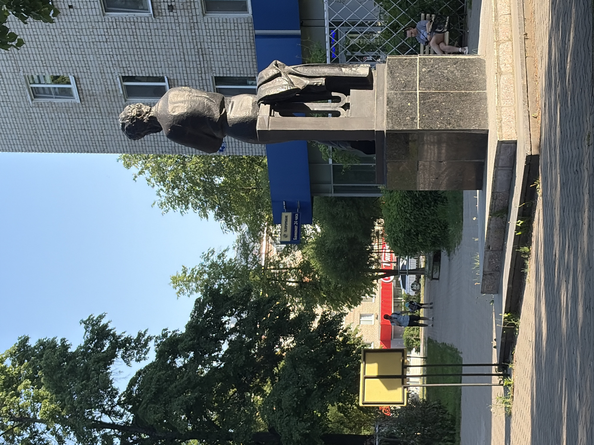 The image captures a street scene in Chaikovsky, Russia. The dominant feature is a bronze statue of a man, positioned in front of a brick building with a blue fascia. A person is seated on the base of the statue.
In the background, the street is lined with trees and a sidewalk. People are walking in the background. Street signage and other urban elements are visible. The day is sunny.