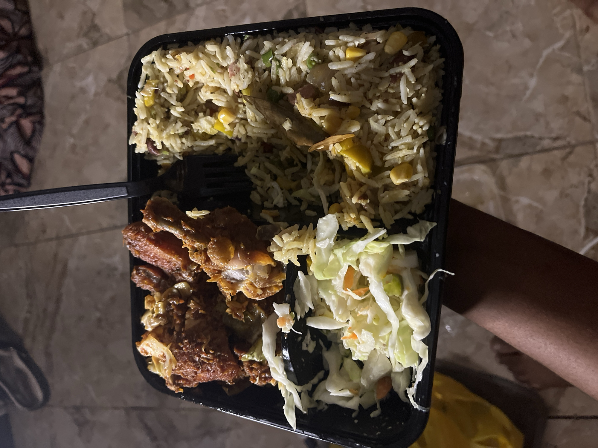 A close-up shot shows a black, partitioned food container held over a patterned floor, possibly marble, and part of a person's arm is visible on the right. The container holds fried rice with corn and peas, fried chicken, and coleslaw. A black plastic fork is partially visible on the left. The environment suggests an indoor setting. There is no indication in the image to determine the exact location in Jalingo, Nigeria.