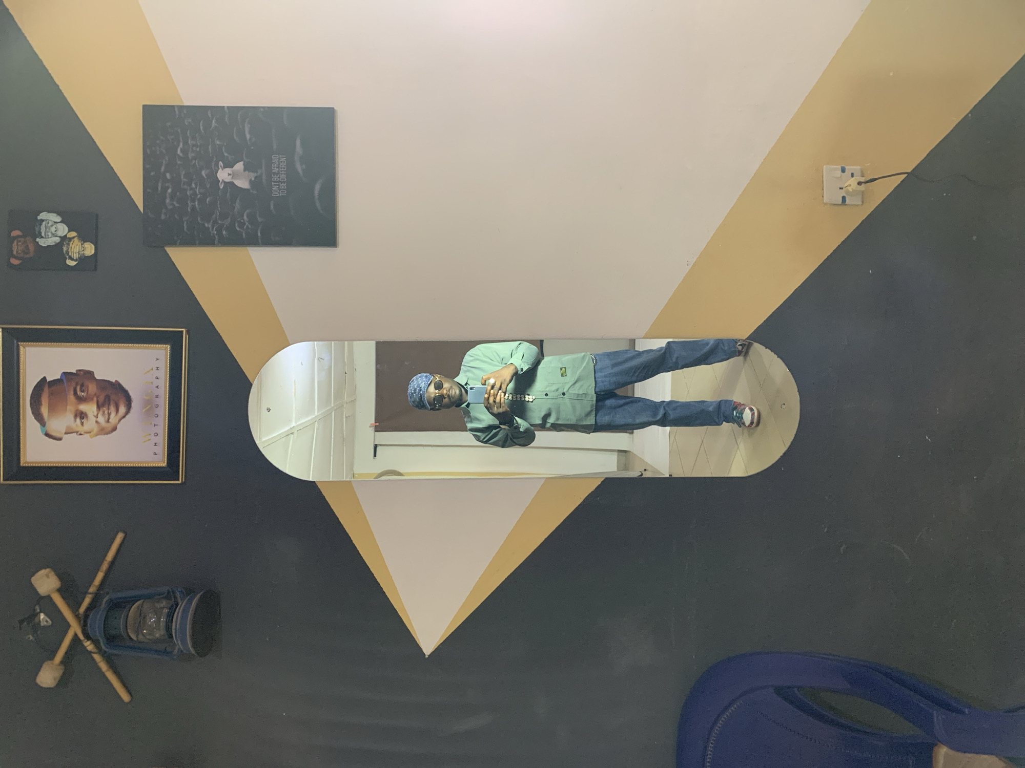 The image captures a man standing in front of a mirror, taking a selfie. He is wearing a light green long-sleeved shirt, blue jeans, a blue patterned headscarf, sunglasses, and white sneakers. The mirror is an oval shape, positioned on a wall with a distinct geometric design featuring black, white, and yellow diagonal stripes. Above the mirror, the wall displays several framed art pieces. On the left, a blue kerosene lamp hangs with crossed wooden sticks above it. To the right of the lamp, a framed portrait of a man is mounted. Above that, a small framed picture depicts three monkeys covering their eyes, ears, and mouth. Below the monkey picture, a larger framed artwork shows a white sheep standing out in a flock of dark sheep, with the text "DON'T BE AFRAID TO BE DIFFERENT" visible. The environment appears to be indoors, possibly a studio or a well-decorated room. A blue chair is partially visible on the left side of the frame. On the right side of the wall, a white electrical socket with a black plug is mounted. This scene is located in Jos, Nigeria, indicated by the presence of a plug socket commonly found in Nigeria.