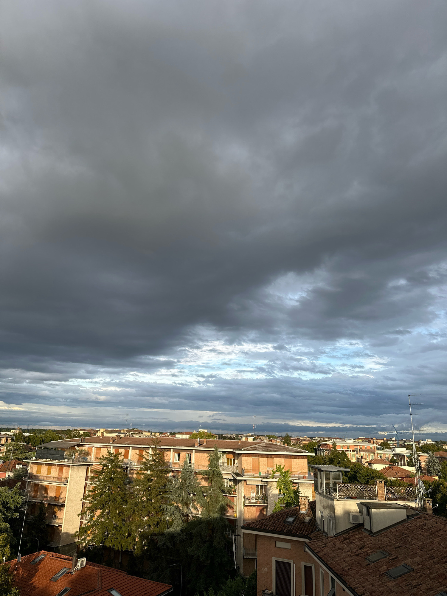 This is a medium-shot, outdoor, horizontal-orientation, eye-level photograph taken from a high vantage point, looking across the rooftops of a city. The sky occupies the top two-thirds of the frame and is filled with a dramatic display of dark, heavy storm clouds. Shafts of sunlight break through in the distance, illuminating sections of the cloud cover with a brighter, blueish hue.
Below the dramatic sky, the city skyline is visible. The dominant features are the red-tiled roofs of various buildings. In the mid-ground, a large, multi-story apartment building with an orange-brick facade and multiple balconies is prominent. There are trees, some with green foliage and others appearing bare or autumnal, interspersed among the buildings. Antennas and small structures are visible on some of the rooftops. The overall impression is of a cloudy, possibly late afternoon or early evening sky over a dense urban or suburban area, likely in Parma, Italy, given the architectural style and the presence of antennas commonly seen on Italian buildings. There are no people or obvious signs of activity depicted.