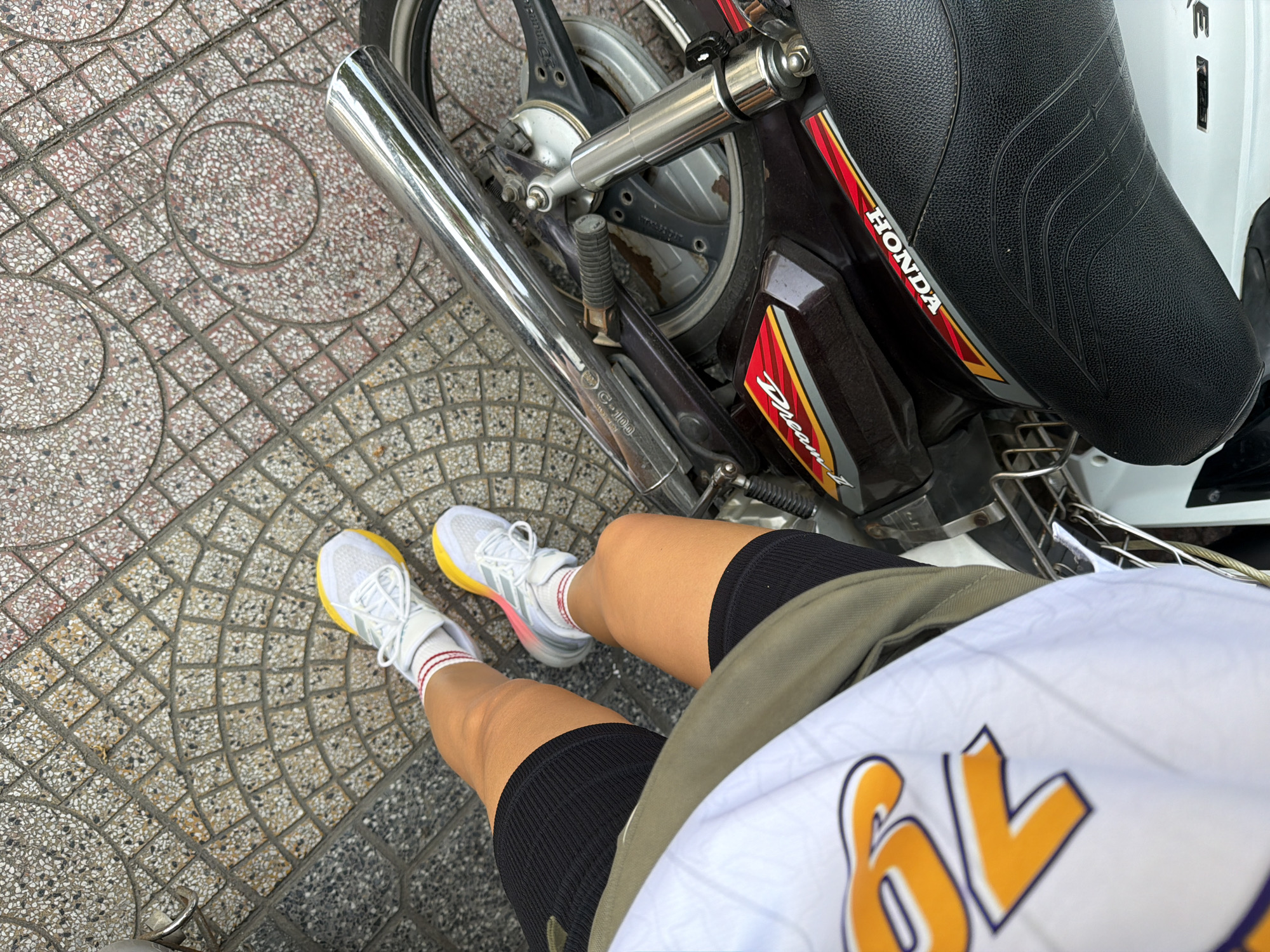 The image shows a person's legs and feet, wearing white athletic shoes with yellow accents and red and white striped socks, resting on the ground next to a dark brown Honda motorcycle. The motorcycle has a chrome exhaust pipe and a black seat. The ground is paved with textured square tiles in a circular pattern. The person is wearing black athletic shorts and a white shirt with orange lettering. The scene appears to be outdoors, on a sidewalk or street in Tay Nha Trang Ward, Vietnam. The lighting suggests it is daytime.