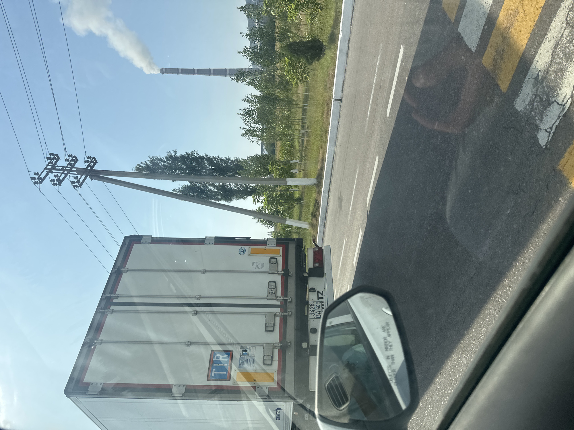 The image depicts a street scene, likely in or near Yanaryk, Uzbekistan, on a sunny day. A large white semi-trailer truck, with the "TR" logo visible on its rear door, is in the foreground, partially obscuring the view of a car's side mirror and the road ahead. The truck has a license plate that reads "3428 BA 40 TZ". In the background, a tall industrial smokestack emits a plume of white smoke against a clear blue sky. There are power lines supported by utility poles, and a line of trees separates the road from the industrial facility. The road has white and yellow markings, suggesting a pedestrian crossing. The overall impression is one of a typical industrial or commercial area on the outskirts of a settlement.