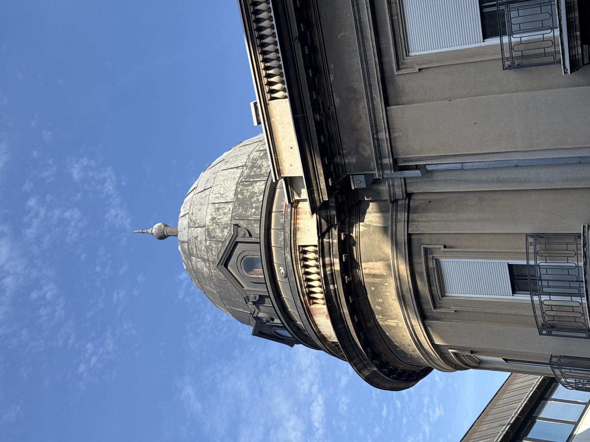 The image is a low-angle shot of a building in Belgrade, Serbia, featuring a dome-shaped architectural element. The building appears to be made of stone or concrete and has windows with white blinds and metal railings. The sky is blue with scattered white clouds.