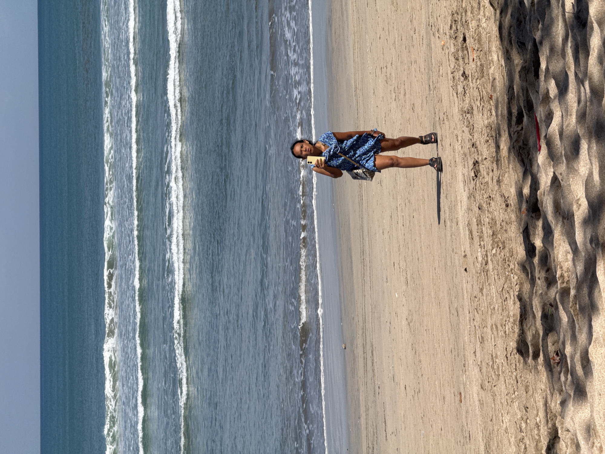 A woman is walking on the beach in Kuta, Indonesia, holding a smartphone in her right hand. She is wearing a blue and white patterned dress and black sandals. A beige bag with a pattern is slung over her left shoulder. The beach is made of light brown sand, with some undulations and shadows from unseen objects in the foreground. In the background, the ocean stretches to the horizon, with multiple lines of breaking waves visible. The sky is a clear, light blue.
