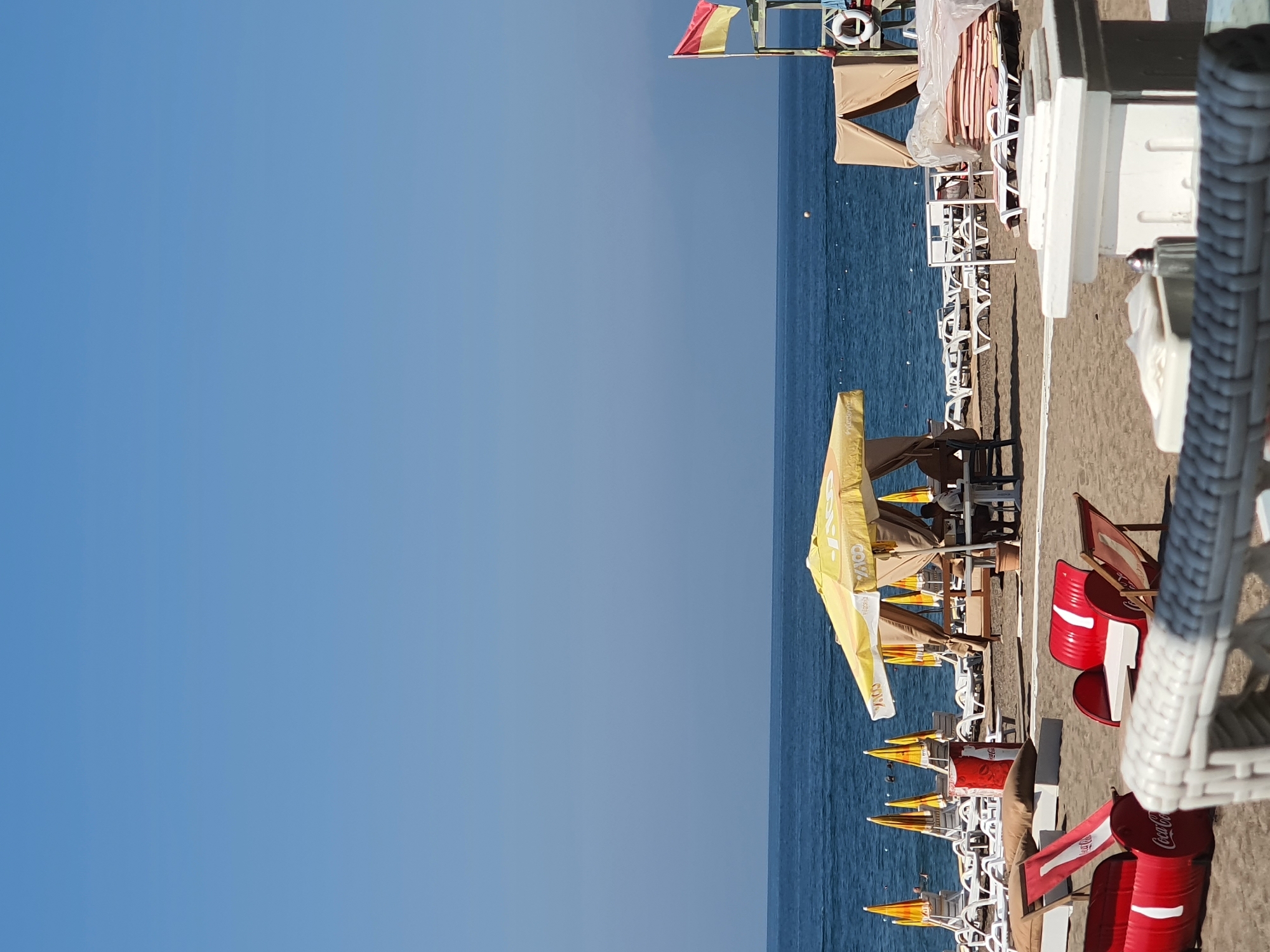 This is a daytime, eye-level, outdoor photograph of a sandy beach in Antalya, Turkey. The scene is dominated by the blue sea and sky. In the foreground, there are several unique, red Coca-Cola branded seating arrangements, made from what appear to be repurposed oil drums. A white, woven chair is also visible in the very front. Behind these, a yellow umbrella shades a seating area with tables and chairs. Further back, rows of white lounge chairs and yellow umbrellas line the beach. The sea is visible in the middle ground, with a red and yellow lifeguard flag visible on a platform to the right. The overall impression is a resort beach setting.