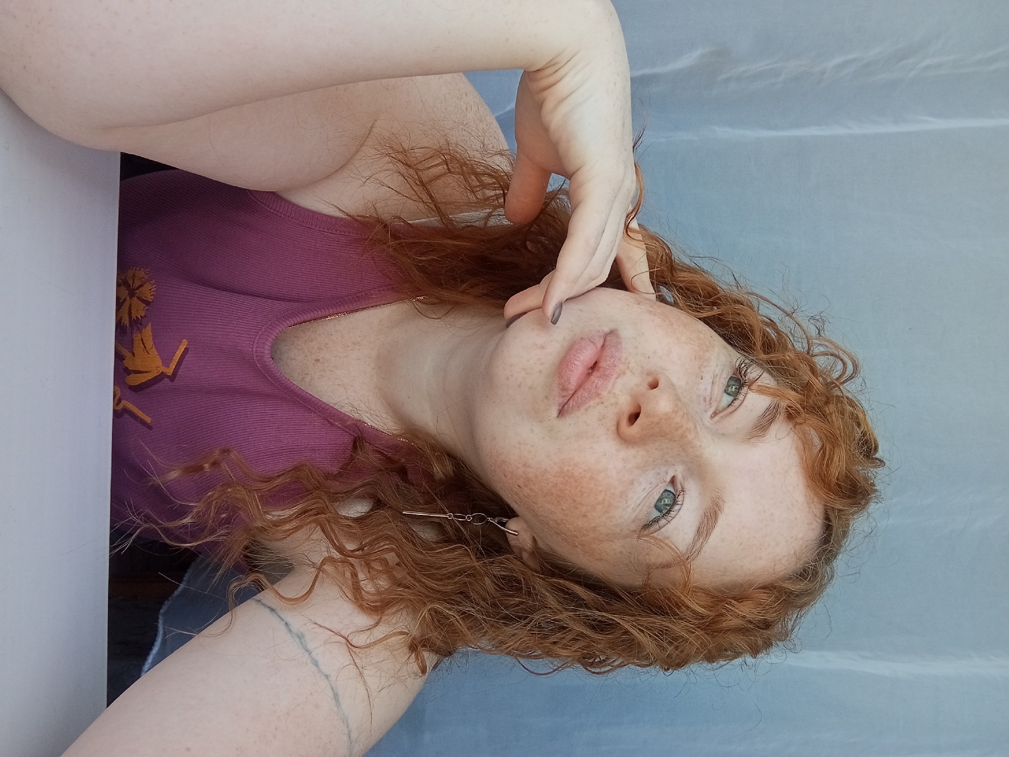 A fair-skinned person with freckles and red curly hair is visible from the chest up. They wear a ribbed, magenta tank top with an orange design on the left side. The person's left hand is raised near their face, with gray-painted fingernails visible. The background is a light blue or gray hue, possibly fabric or a wall. An earring is visible on the lower left. A tattoo is present on the arm in the lower left of the image. The image is rotated, with the subject appearing sideways. The location is not indicated in the image and is therefore indeterminable.