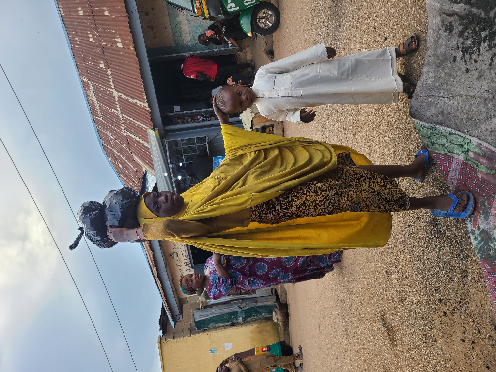 In the city of Jos, Nigeria, on an overcast day, a young girl, possibly in her early teens, carries a load of black trash bags balanced on her head. She is wearing a bright yellow hijab and a patterned skirt. To her right, a young boy in a cream-colored tunic stands, looking towards the viewer. In the background, an older woman in a patterned dress and headscarf stands near a shop with a corrugated metal roof. Another person in a brown outfit is visible to the far left, standing near a three-wheeled vehicle. The scene appears to be a street or market area, with a dusty, paved ground and buildings lining the street. The overall impression is one of everyday life in a bustling environment.