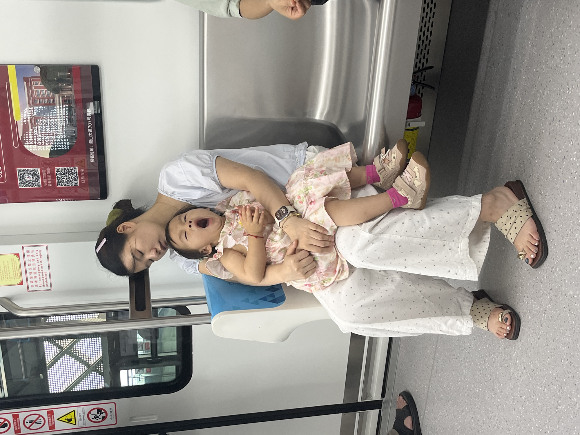 A woman is sitting on a subway train in Tiaodeng, China, holding a young child on her lap. The woman is wearing a white shirt and white pants with a floral pattern. She has her head tilted back, with her eyes closed. The child is wearing a pink floral dress and has their mouth wide open, appearing to yawn. The child is also wearing pink socks and light-colored sneakers. The woman is wearing sandals with a polka-dot pattern. The subway car appears to be clean and modern, with metal seats and white walls. There are signs on the wall, some of which are in Chinese. The floor of the subway car is a light gray speckled material.