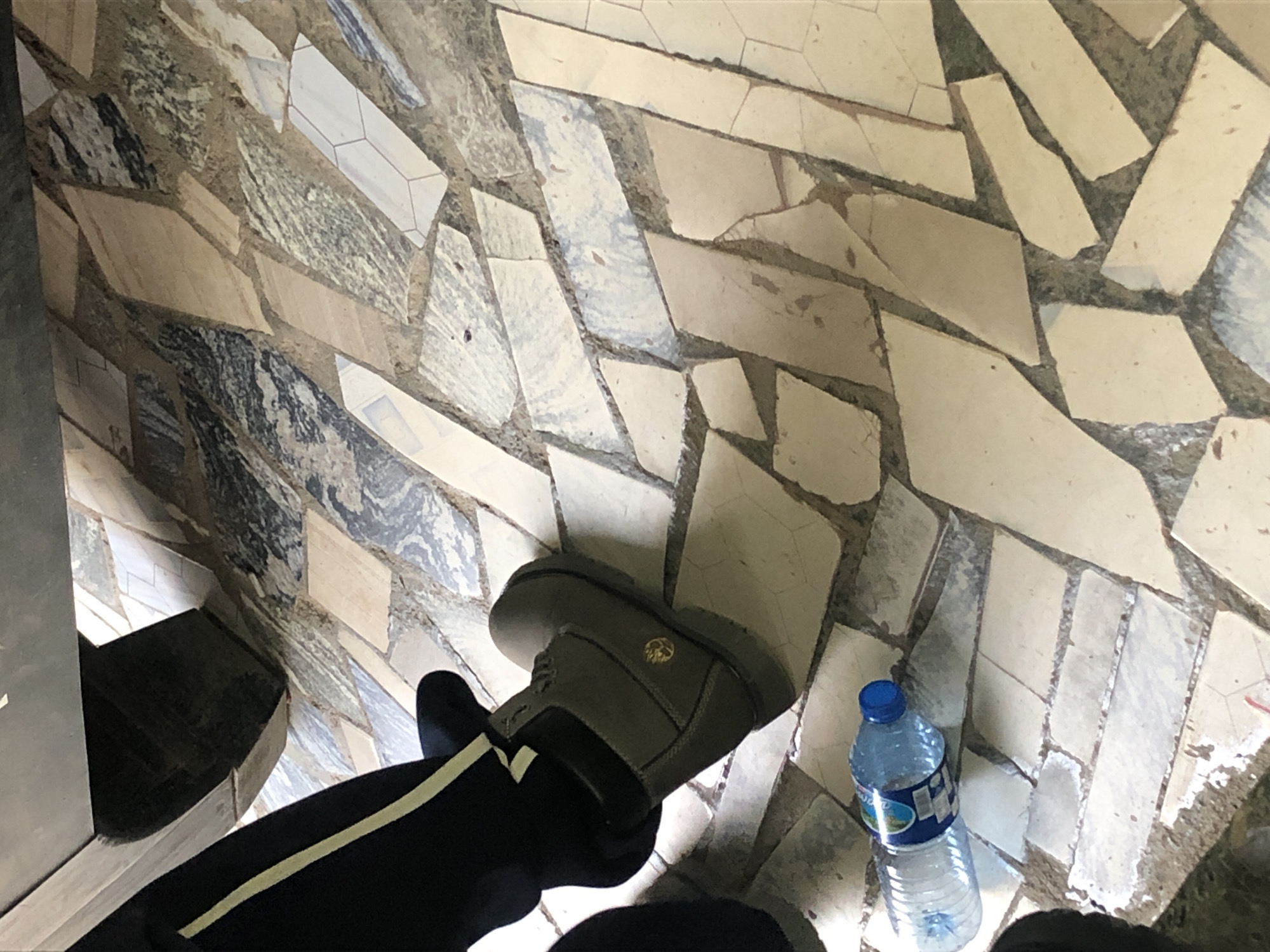 A low-angle, close-up shot shows a person's lower leg and foot, clad in black pants with a yellow stripe, and a grey Timberland boot. The boot rests on a floor tiled with irregular, flat pieces of stone, predominantly in light beige and grey tones, interspersed with darker grey, black, and white marbled stones. The grout between the tiles is a sandy, light brown color. To the left of the boot, a clear plastic water bottle with a blue cap and a partially visible label lies on the floor. The background is dark, suggesting a shadowed area or an opening. The scene is outdoors on a patterned stone floor.