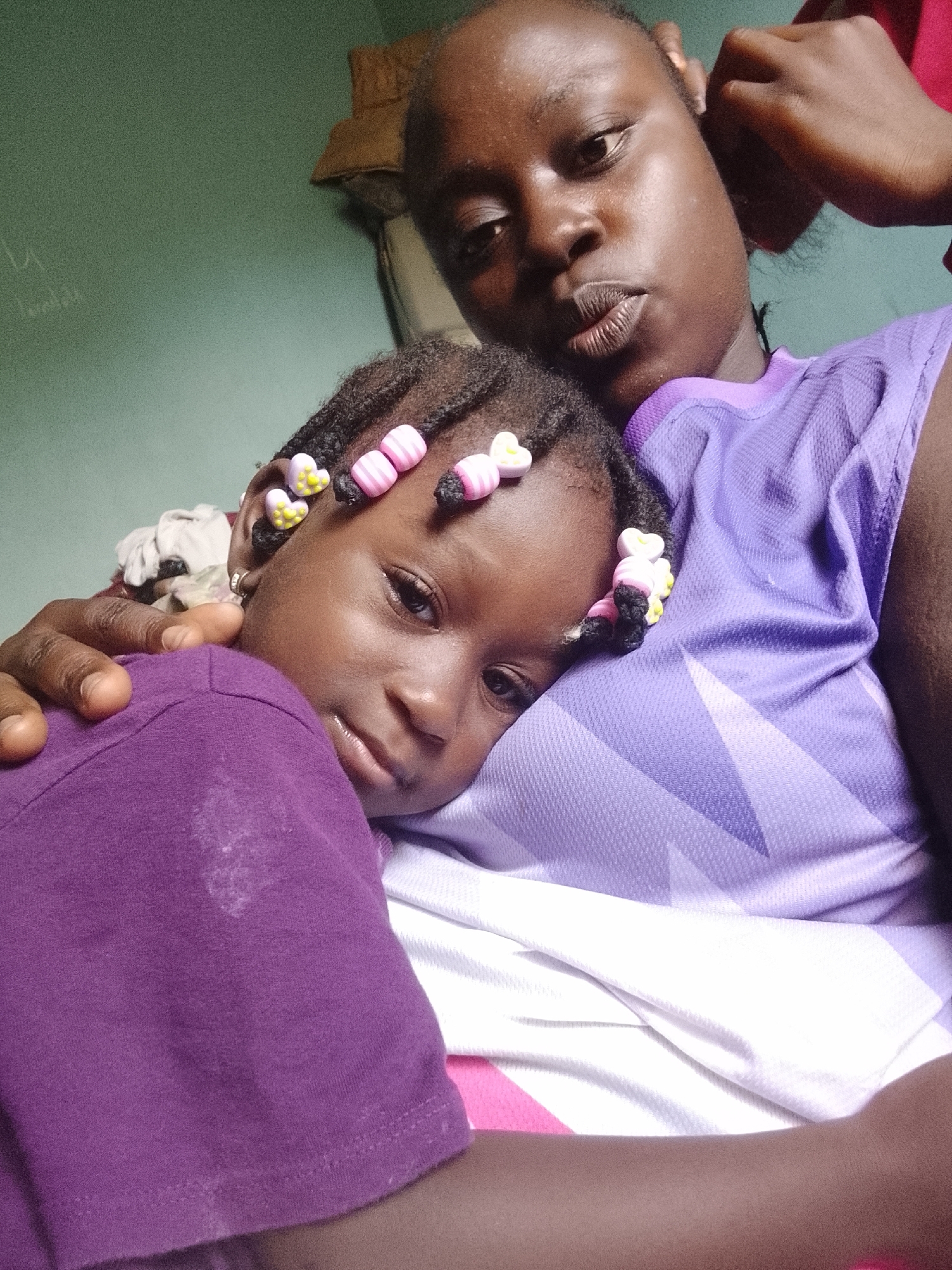A medium shot depicts a young woman holding a child. The woman has dark skin, short dark hair, and is wearing a purple t-shirt. The child is dark-skinned and has braided hair adorned with pink and yellow beads. The child is wearing a purple t-shirt and is being held in the woman's arms. The background includes a light green wall and some indiscernible objects. The location is Unknown, Unknown.