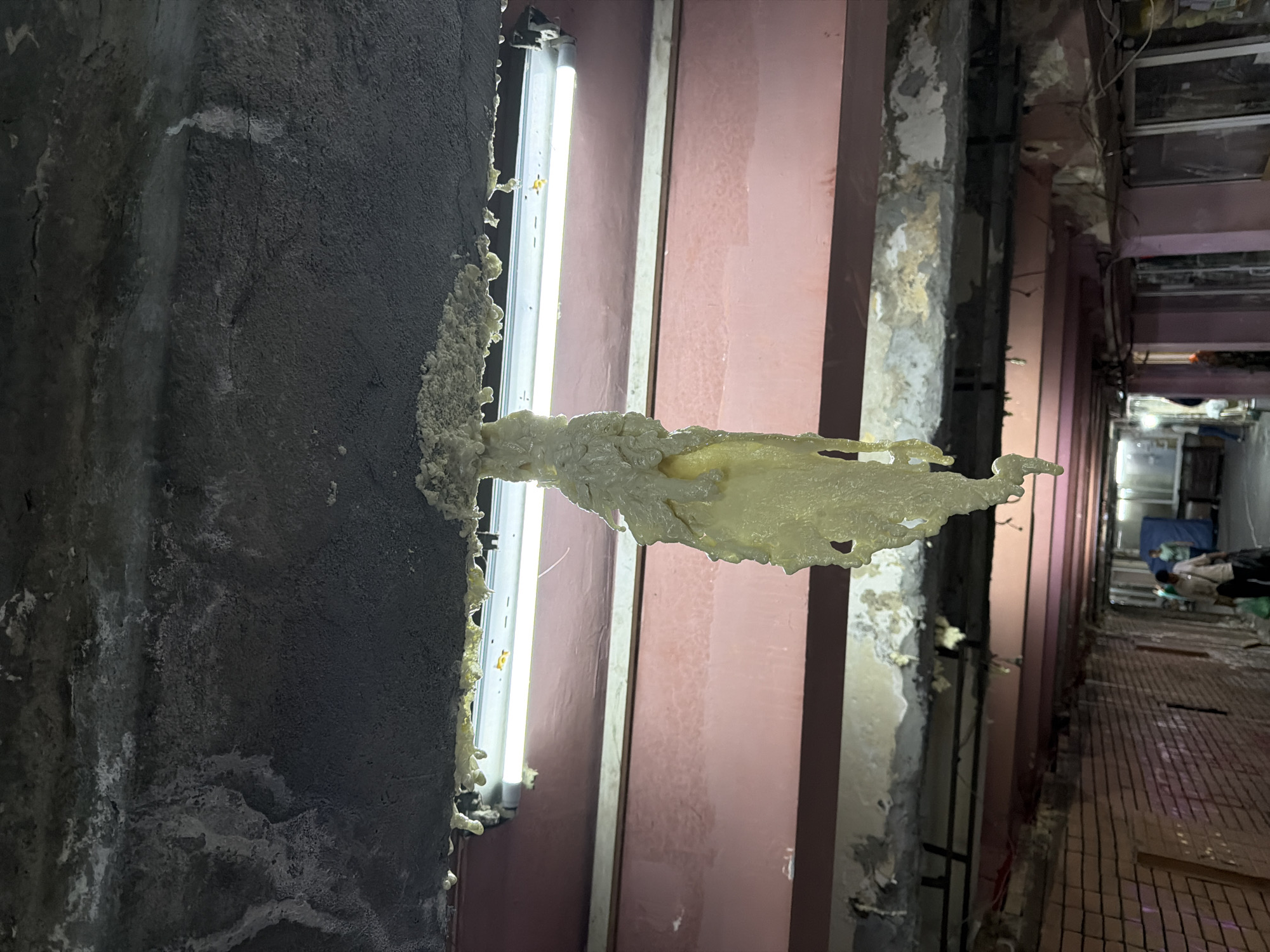 The image depicts an underpass or tunnel-like structure, likely in Tbilisi, Georgia, exhibiting signs of disrepair. A prominent feature is a thick, yellow, foam-like substance dripping from the ceiling, forming a large, irregular mass. The ceiling itself appears to be concrete, with visible cracking and some lighter-colored efflorescence or mineral deposits. A fluorescent light fixture is mounted horizontally on the ceiling, directly above where the foam is dripping.
Below the ceiling, there is a large pink beam, also showing some signs of wear. Further down, another level of beams, possibly supporting the structure, is visible. The walls of the tunnel are lined with reddish-brown brickwork on the left side and appear to be painted a pinkish color on the right. Wires and conduits are visible along the upper parts of the right-hand wall.
In the background, several individuals are present, walking through the tunnel. One person in the foreground on the right is carrying plastic bags. The overall environment suggests an urban passage that is either undergoing renovation, suffering from neglect, or has experienced some form of damage, possibly water-related, given the dripping foam and ceiling degradation. The lighting is artificial, provided by the fluorescent fixture, and the scene is captured in a downward perspective, emphasizing the dripping substance.