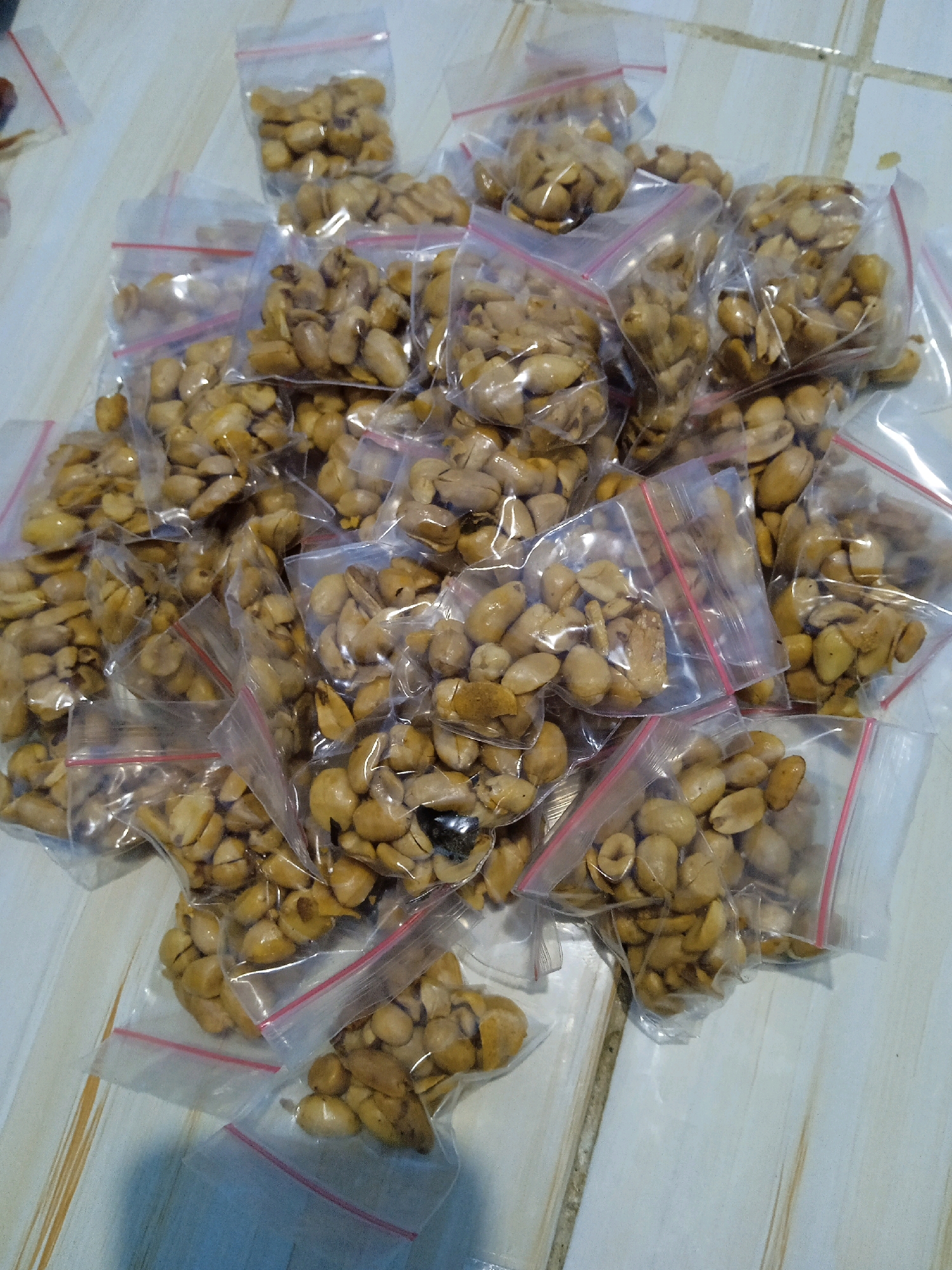 A pile of small, clear plastic bags containing roasted peanuts is scattered across a light-colored tiled surface. The bags are mostly filled with the nuts and appear to have red zippers. The image is taken from a top-down perspective. The scene is likely a market stall or home-based food preparation area in Banjarmasin, Indonesia, given the common practice of selling food in such small, individual portions. There are no people or animals visible in the image. The lighting is overhead and casts minimal shadows, suggesting an indoor setting.