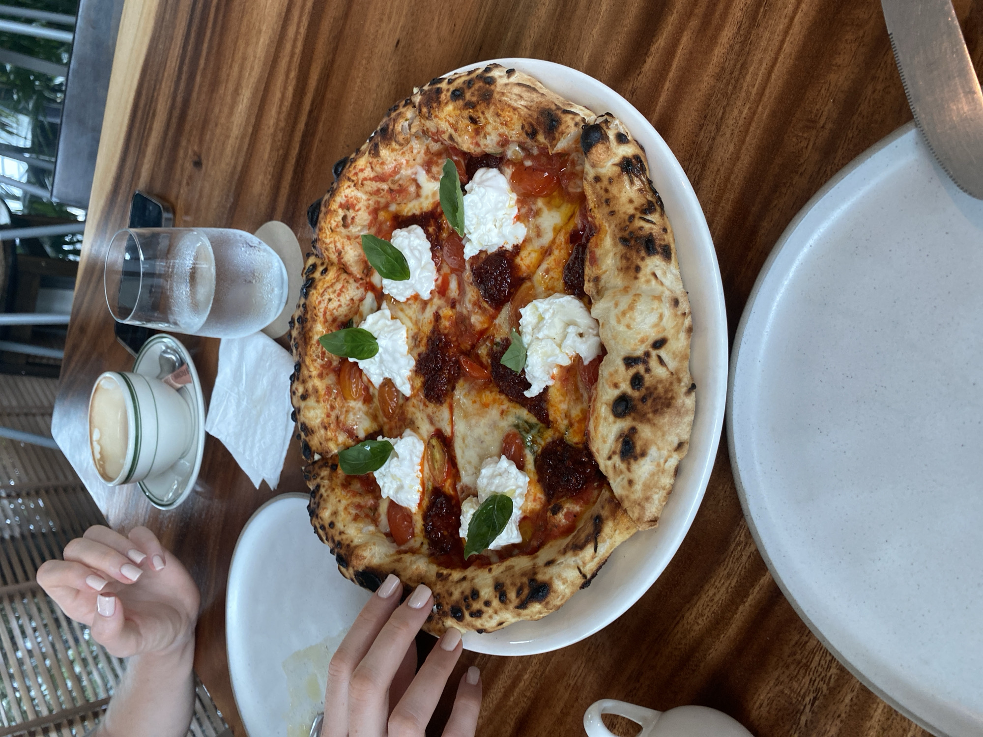 A pizza is centered in the image on a white plate, sitting on a dark wooden table. The pizza has a thick, charred crust with toppings of tomato sauce, white cheese (possibly burrata), and fresh basil leaves. To the left of the pizza is a white teacup with a cappuccino or latte on a saucer, a glass of water, and a person's hand with light skin and painted nails reaching towards the pizza. To the right of the pizza is a white plate with a knife on it. The setting appears to be a restaurant in Choeng Thale, Thailand.