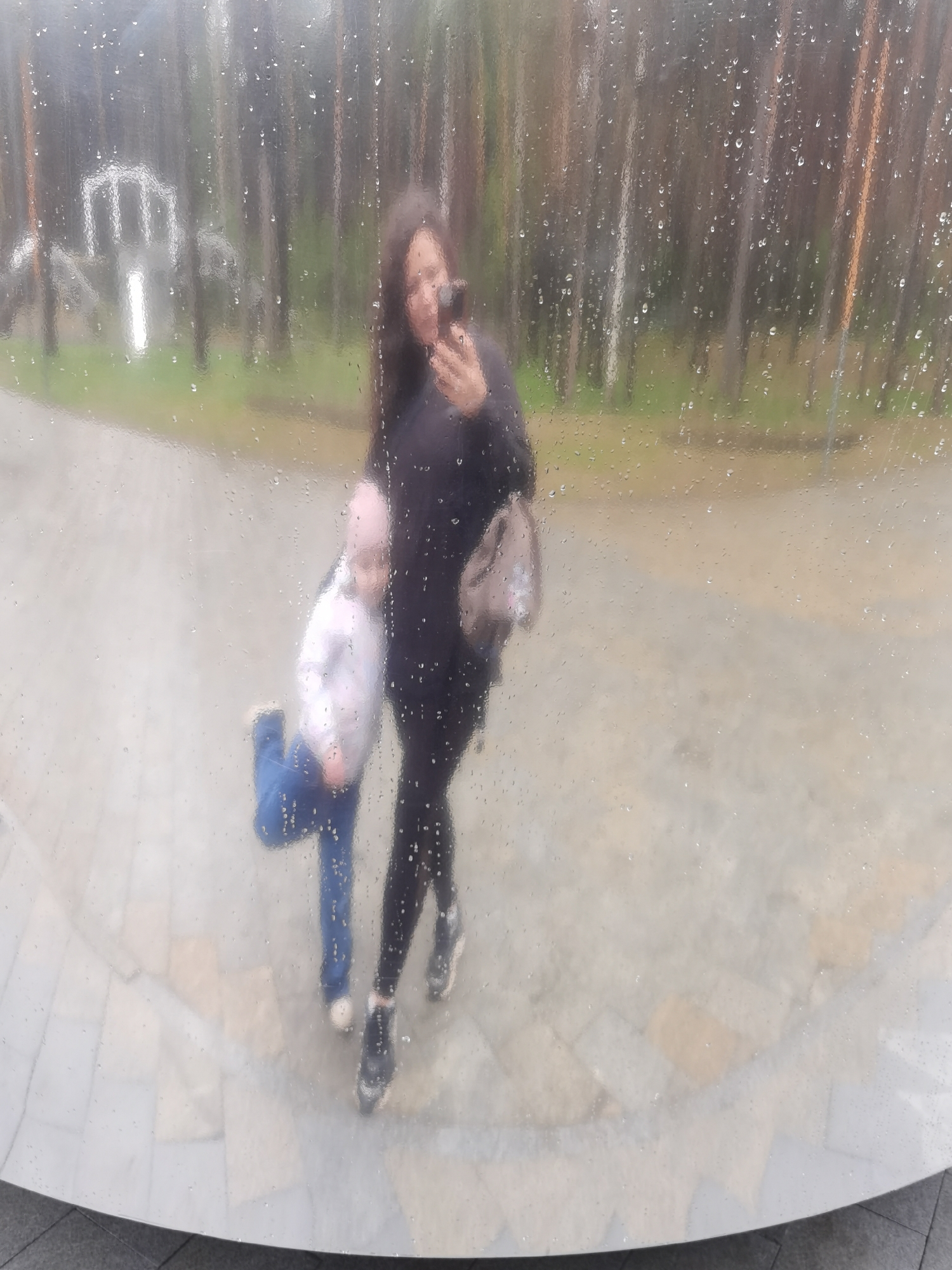 The image shows a reflection of a woman taking a selfie with a child in front of a mirrored sculpture. The reflection is obscured by raindrops on the surface. The woman is wearing a black top and leggings, sneakers, and carrying a purse. The child is wearing a light colored top and blue pants. In the background, trees and an architectural structure are visible through the reflection. The ground shows pavement and decorative stone elements. Based on the context clues of the sculpture and background, this is likely a photograph taken at the “Permyak Salty Ears” sculpture in the city of Perm, Russia.