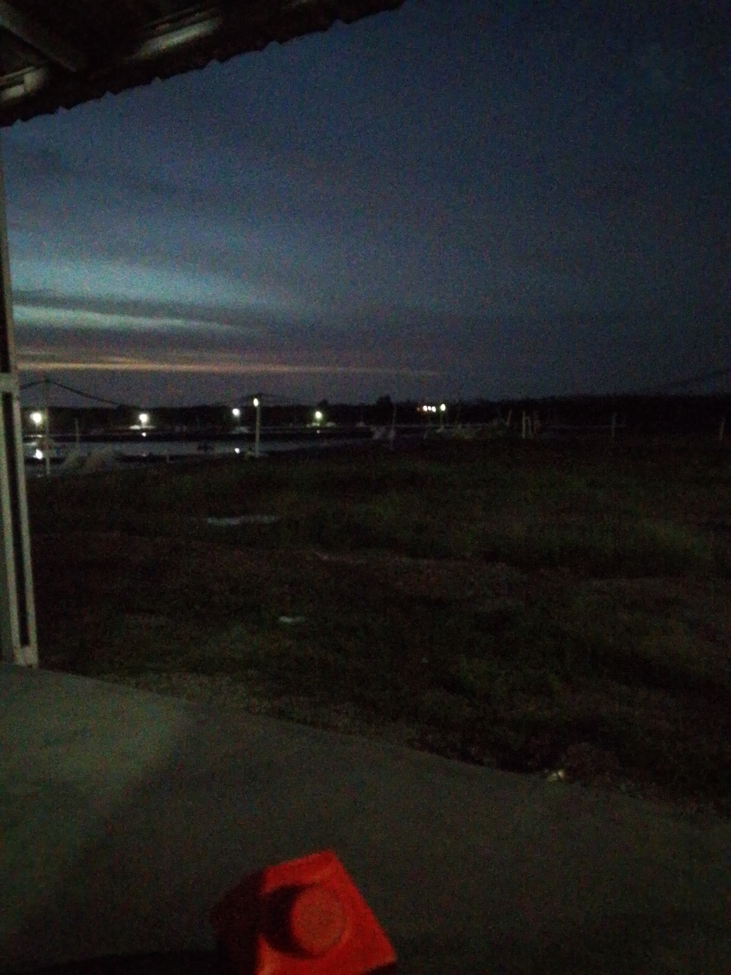 The image shows an outdoor scene in Bukit Ketok, Indonesia at dusk or night. In the foreground, there is a part of a building structure, including a portion of a roof and a white pillar. On the ground in the lower portion of the image, there is a red plastic object with a circular top. Beyond the foreground, there is a grassy field and a body of water. Several lights are visible across the water in the distance. The sky is dark with hints of lighter colors near the horizon, suggesting a sunset or twilight. The overall lighting is low, making it difficult to discern fine details.