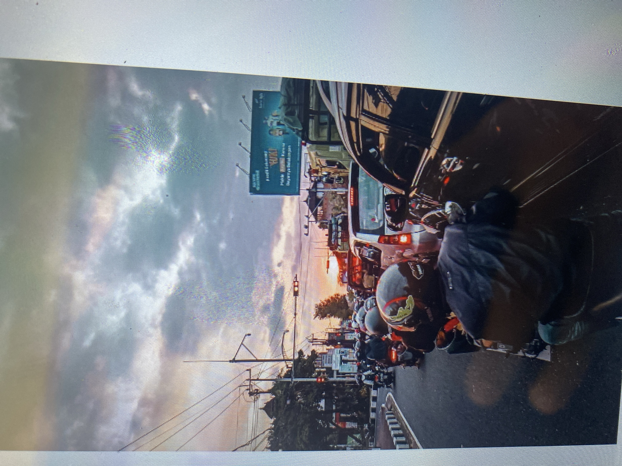 This image depicts a congested street scene in Ta', Indonesia, during sunset. The sky is filled with dramatic clouds, colored by the setting sun. A long line of motorcycles and cars are stationary on the road, indicating heavy traffic. Several individuals are visible on motorcycles, wearing helmets, suggesting they are commuters. In the background, there are buildings, trees, and a large billboard advertisement. Traffic lights are visible, some appearing red, further suggesting a traffic stop. The overall environment appears to be an urban setting with overhead power lines and street infrastructure.