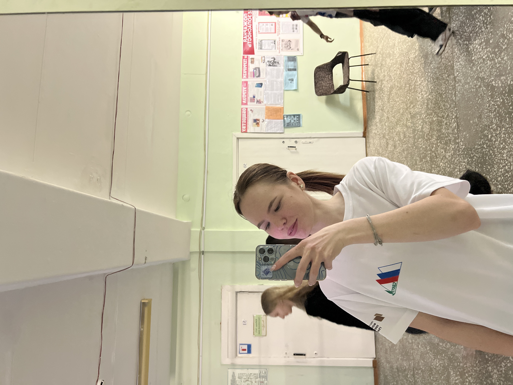 A young woman is taking a selfie in front of a mirror inside a public space, likely a school or community center, based on the presence of informational posters and a notice board on the wall. The environment includes a tiled floor, a chair, and a door. The reflection in the mirror shows a person walking in the background. The woman is wearing a white t-shirt with a Russian flag logo. The scene is likely in Berezniki, Russia, because the Cyrillic text on the informational posters and on the woman's t-shirt implies a Russian location.