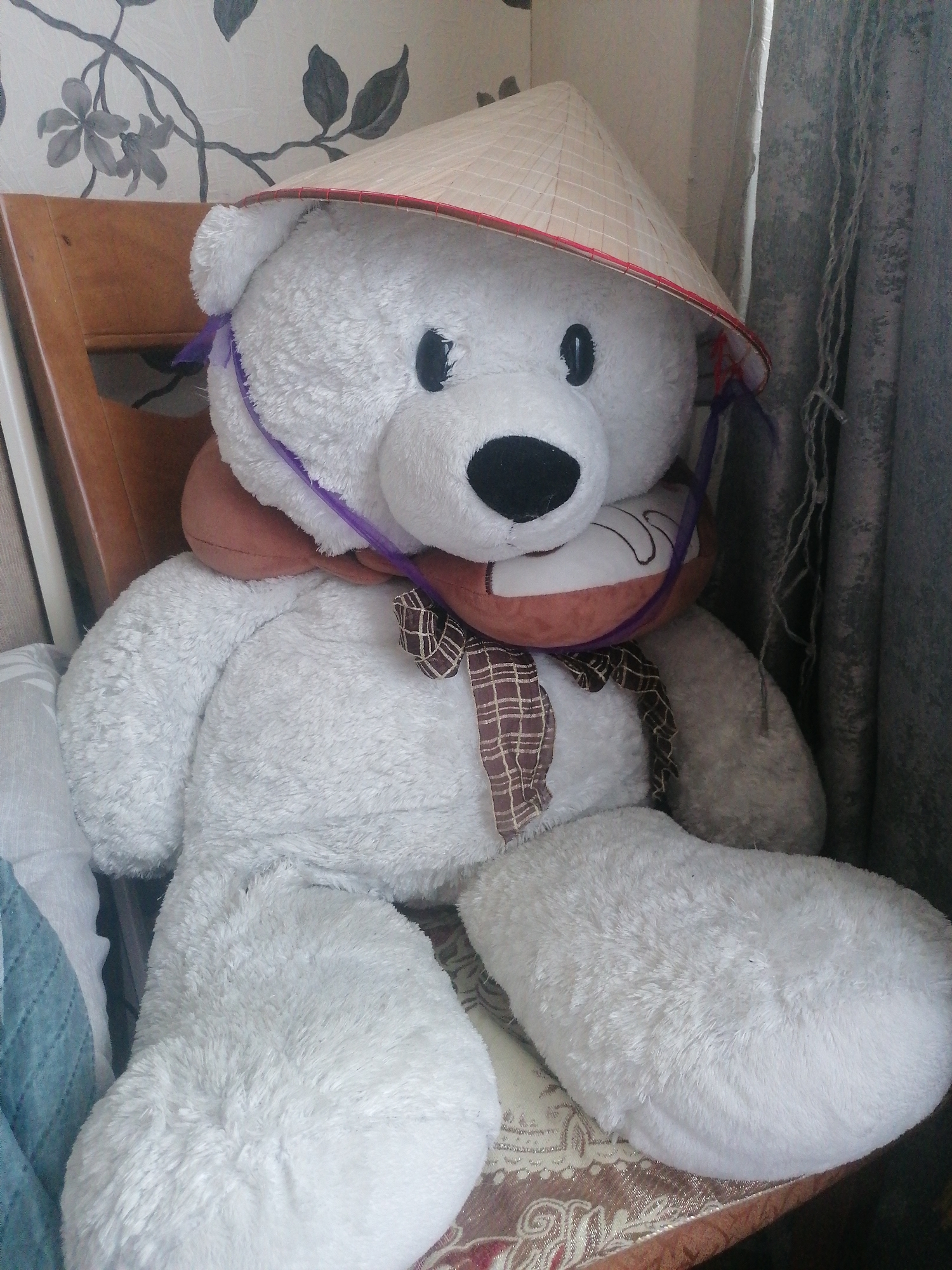 A large, white teddy bear is seated on a wooden chair, indoors. The bear is wearing a conical, woven Asian hat, a brown and plaid scarf around its neck, and a purple ribbon tied around its head under the hat. The bear's face has black eyes and a black nose. The background consists of a floral patterned wallpaper on the left and grey curtains on the right. A patterned fabric is visible beneath the bear. The scene is lit by ambient indoor lighting. There are no people present. The location is identified as Angarsk, Russia.