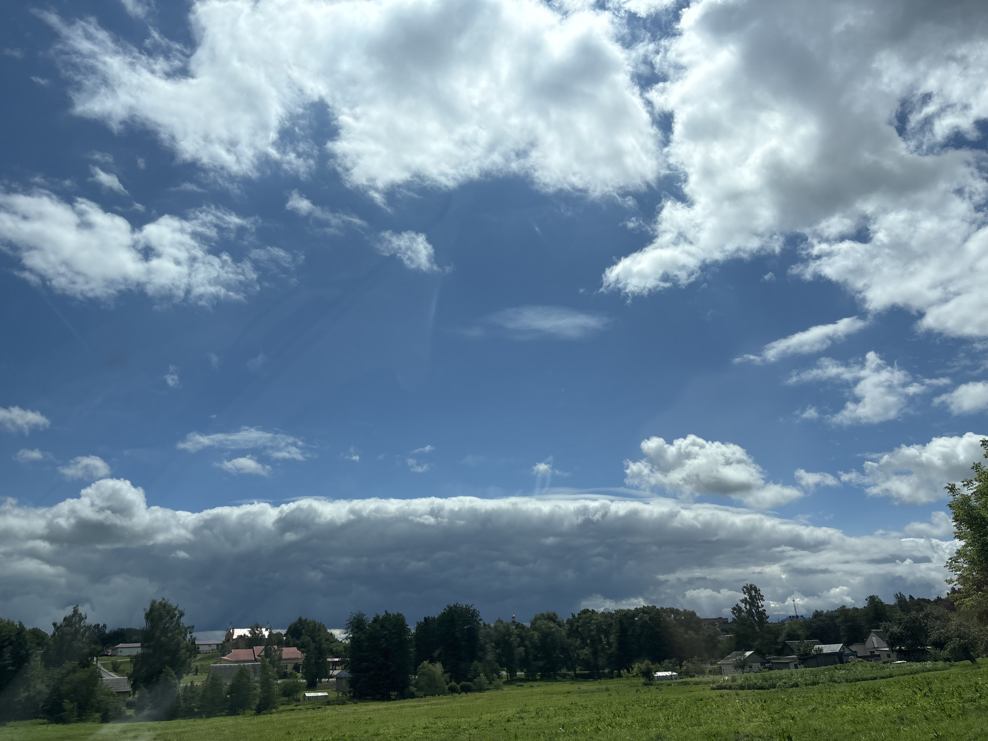 The image shows a view of the Капыльскі сельскі Савет, Belarus, with a vast blue sky filled with dramatic clouds. In the foreground, a lush green meadow stretches towards a line of trees and scattered houses with red roofs. A long, dark, horizontal cloud formation dominates the middle ground, casting a shadow over the landscape below. Above this, wispy white clouds drift across the bright blue sky, with rays of sunlight visible through some of them. The scene appears to be a rural landscape on a partly cloudy day.