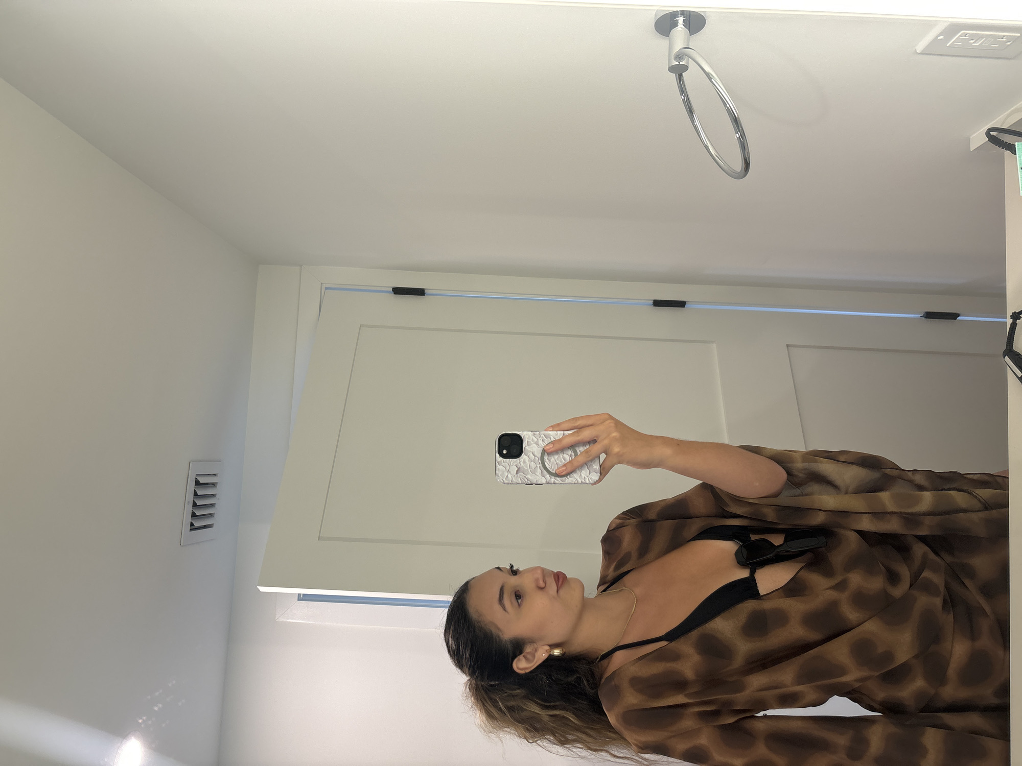 A young woman is taking a mirror selfie in a bathroom in Hallandale Beach, United States of America. She has her hair in a high ponytail and is wearing a black bikini top and a brown, patterned cover-up. She is holding a smartphone in her right hand. In the background, there is a closed white door and a silver towel ring mounted on a white wall. A white air vent is visible on the ceiling.