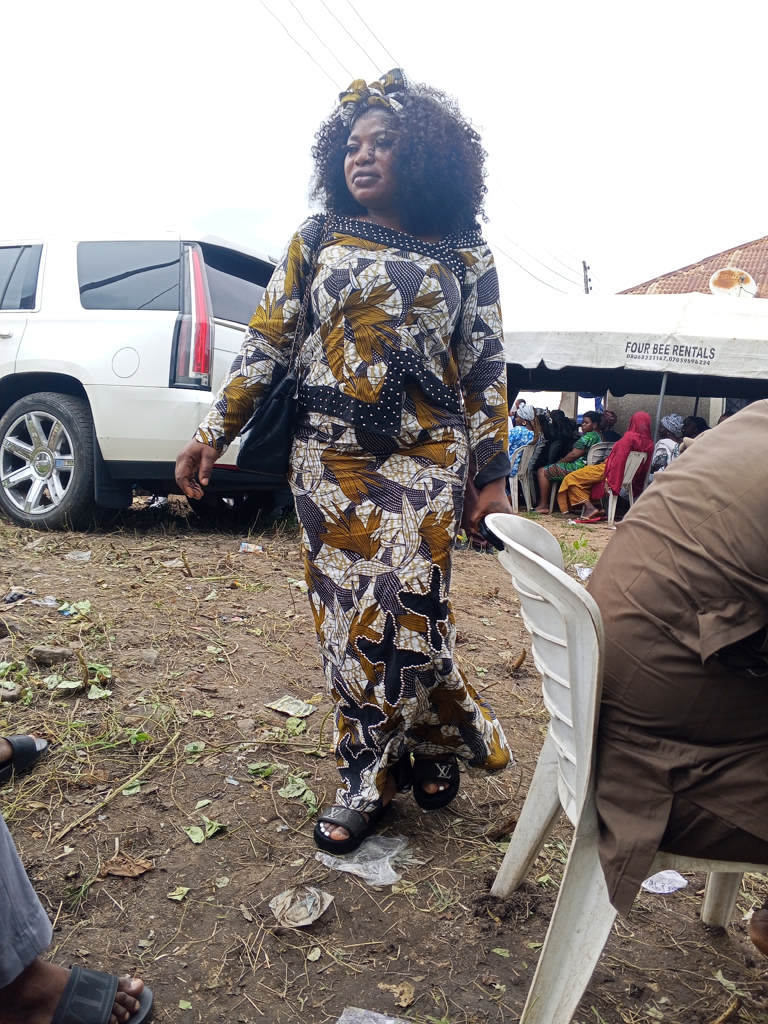 A woman in a patterned dress walks across a dirt lot in Lokoja, Nigeria, near a white SUV and a tent with "FOUR BEE RENTALS" printed on it. She has dark, curly hair and is wearing a headscarf. Several people are seated under the tent in the background. The ground is covered with dry grass, leaves, and some litter. The sky is overcast.