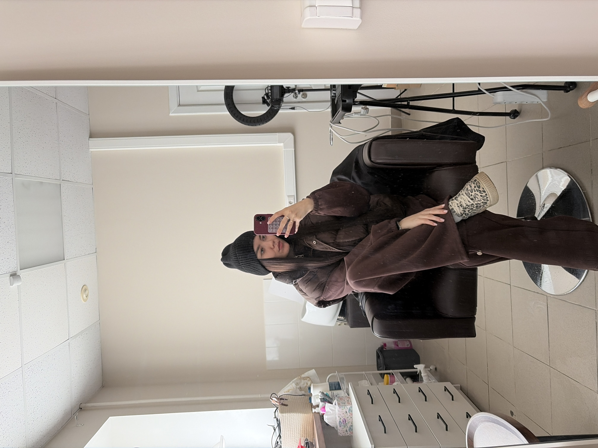 This indoor mirror selfie captures a young woman seated in a dark brown salon chair within what appears to be a beauty or hair salon. The image is taken as a reflection, showing the woman holding a dark-colored smartphone to capture herself. She is dressed casually in a dark beanie, a brown puffer jacket, and brown wide-leg pants, paired with distinctive leopard print high-top sneakers. Rings adorn her fingers.
The setting features light-colored walls and a white dropped ceiling with embedded lighting panels. Behind the woman in the reflection, a ring light on a stand is visible, along with various power cables and a power strip, suggesting a setup for photography or video. To her right, a white cabinet or dresser with several black pull handles holds an assortment of salon supplies including bottles, a tissue box, and a woven basket. A white ceramic basin or sink is partially visible in the background. The floor appears to be tiled. The overall ambiance is clean and functional, typical of a commercial establishment. There is no visible text. The activity is a casual mirror selfie, likely taken in Chaikovsky, Russia.