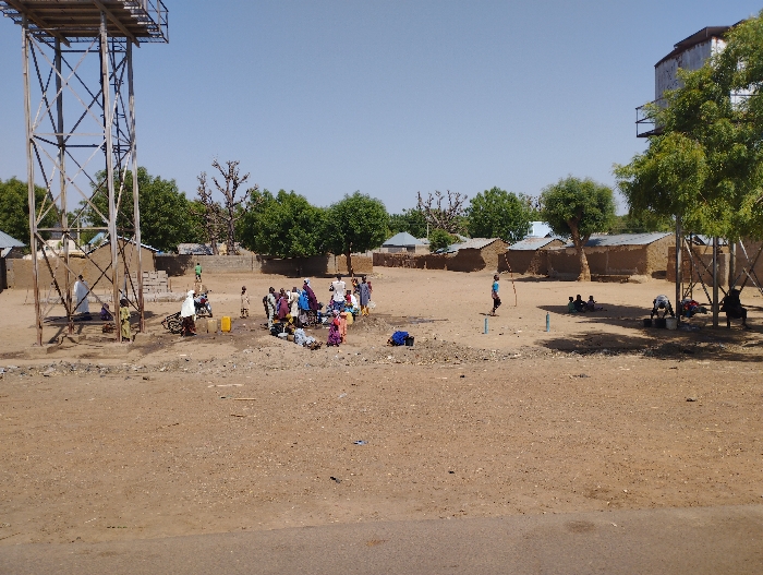 The image shows a scene in Tongo, Nigeria, with a large water tower in the foreground and a smaller water tower in the background. Several people, mostly children, are gathered in a dirt area in front of the larger water tower. Some are sitting on the ground, while others are standing. There is a motorbike in the background. The scene appears to be a gathering place for the community. The ground is dry and sandy, and there are several trees in the background. The houses in the background are simple, made of mud bricks. The sky is clear and blue.