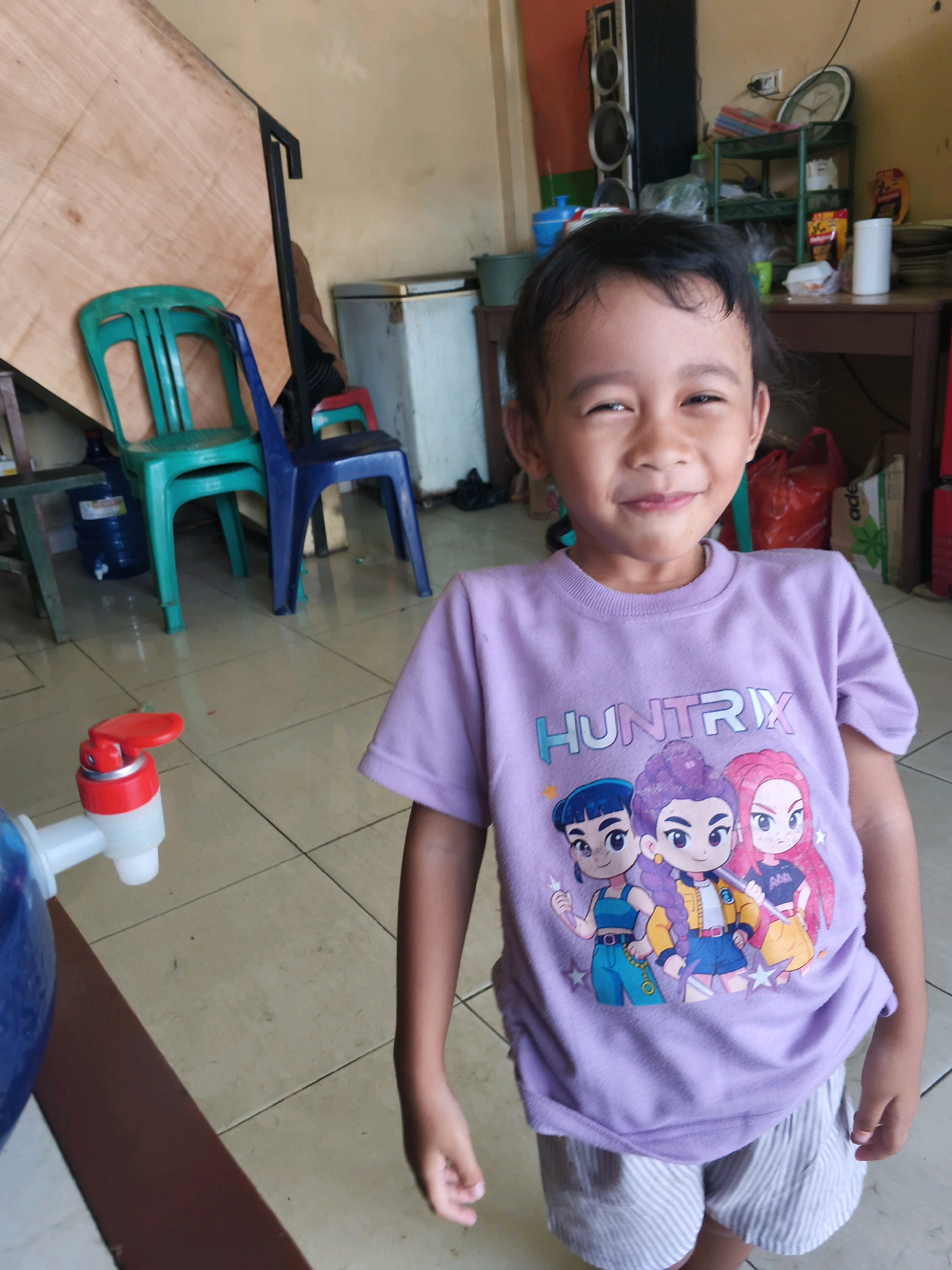 A young Indonesian child, estimated to be between 4 and 6 years old, stands in a room with a tiled floor. The child is wearing a purple t-shirt with an animated design featuring three characters and the word "HUNTRIX", and light-colored striped shorts. The child is smiling and looking at the camera. In the background, several objects are visible, including a large sheet of wood leaning against a wall, a green plastic chair, a blue plastic chair, a white refrigerator, a water dispenser with a red tap, a speaker, shelves with various items, and red plastic bags. The setting appears to be a residential or storage area in Sukoharjo III, Indonesia.
