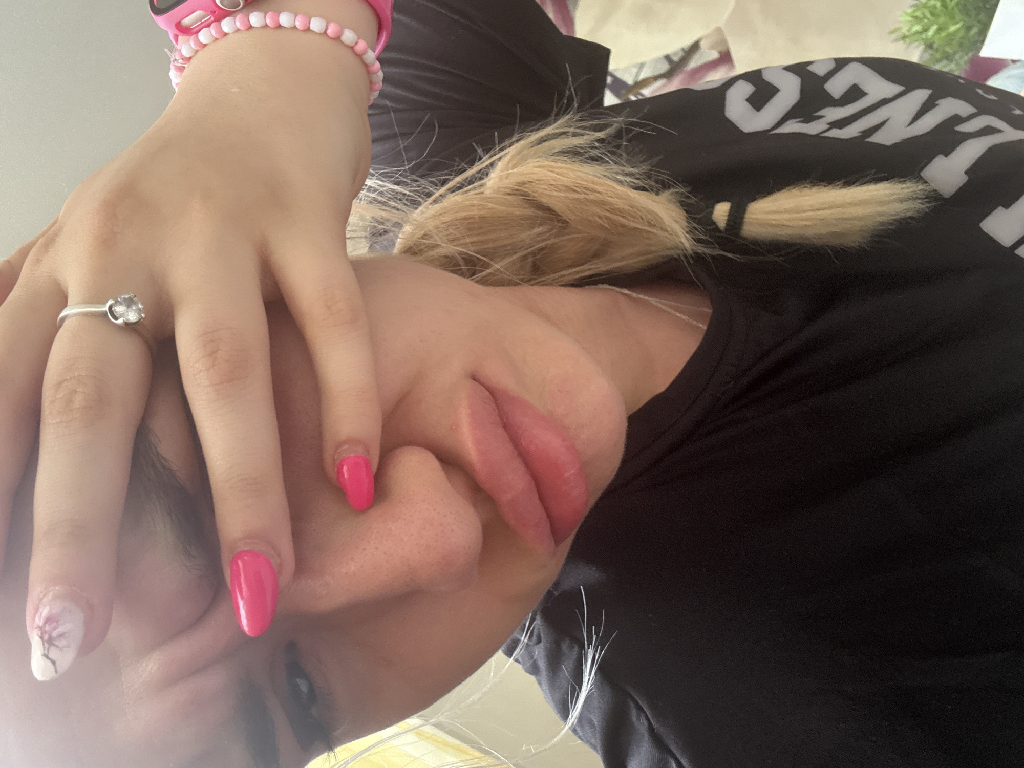 A selfie taken from a low angle features a woman's face with blonde hair, partially obscured by her hand. The hand has an assortment of colorful nails, and a silver ring. She is wearing a black T-shirt with white lettering. A pink bracelet is visible on her wrist. The environment is blurry and difficult to discern. There is no information available to indicate the location is Sofia, Bulgaria.