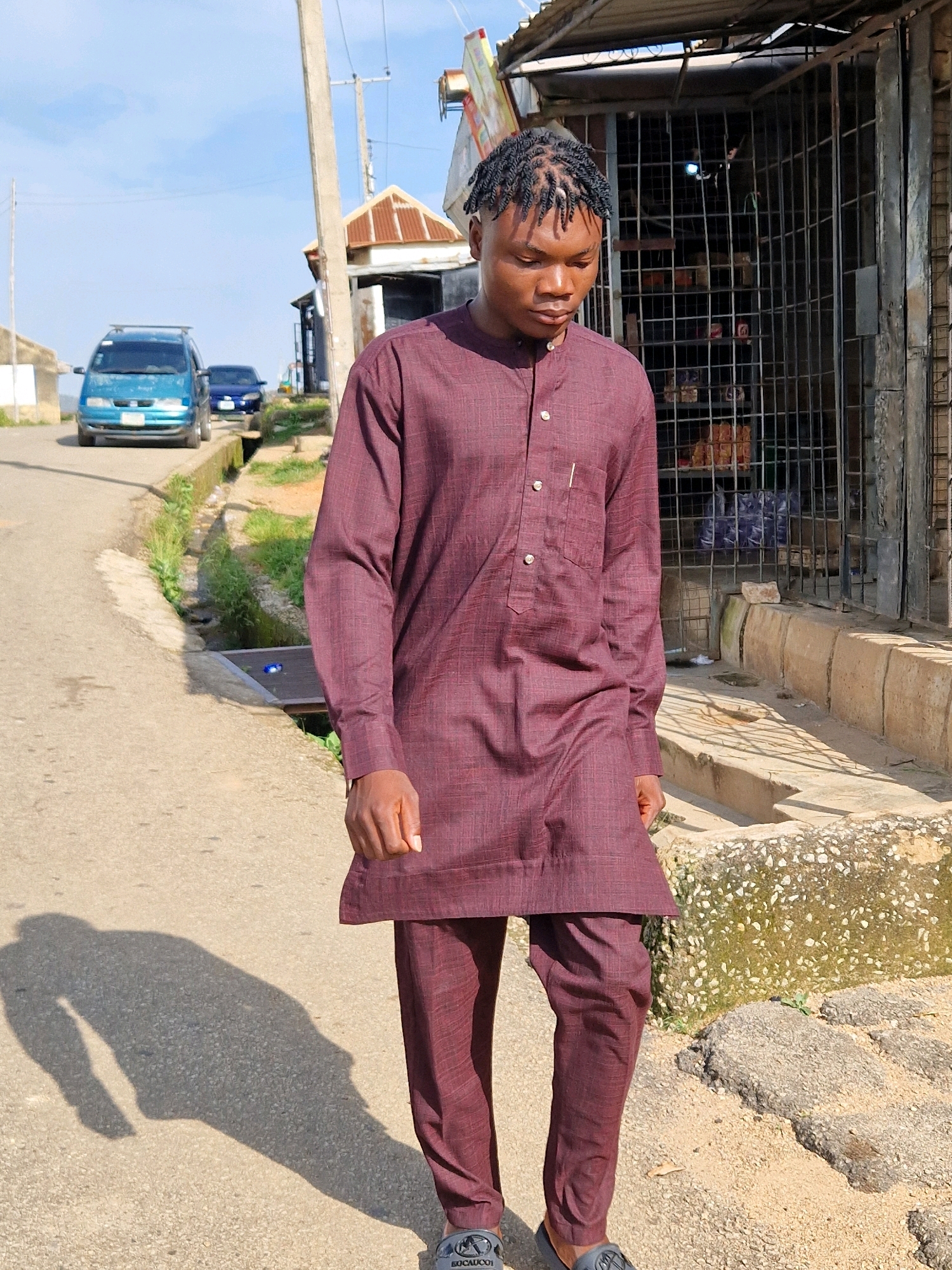 A young Black male with dark, braided hair stands on an asphalt road in Jos, Nigeria, facing slightly away from the camera and looking down. He wears a maroon two-piece outfit consisting of a long-sleeved tunic with a Mandarin collar and two buttons, and matching trousers. The tunic has a single breast pocket on the left side. His right hand is visible at his side, and his left hand is obscured. His shadow stretches to his left on the road.
The road is bordered by a concrete drainage channel on his right, which contains some greenery and a metal grate. Beyond the drainage channel, a metal gate encloses a structure that appears to be a shop, with shelves visible inside. A blue minivan and a dark blue car are parked on the road in the background to the left, with other structures and vegetation also present. The sky is clear with some clouds, and the lighting suggests it is daytime. A tall, grey utility pole is visible to the left of the subject.