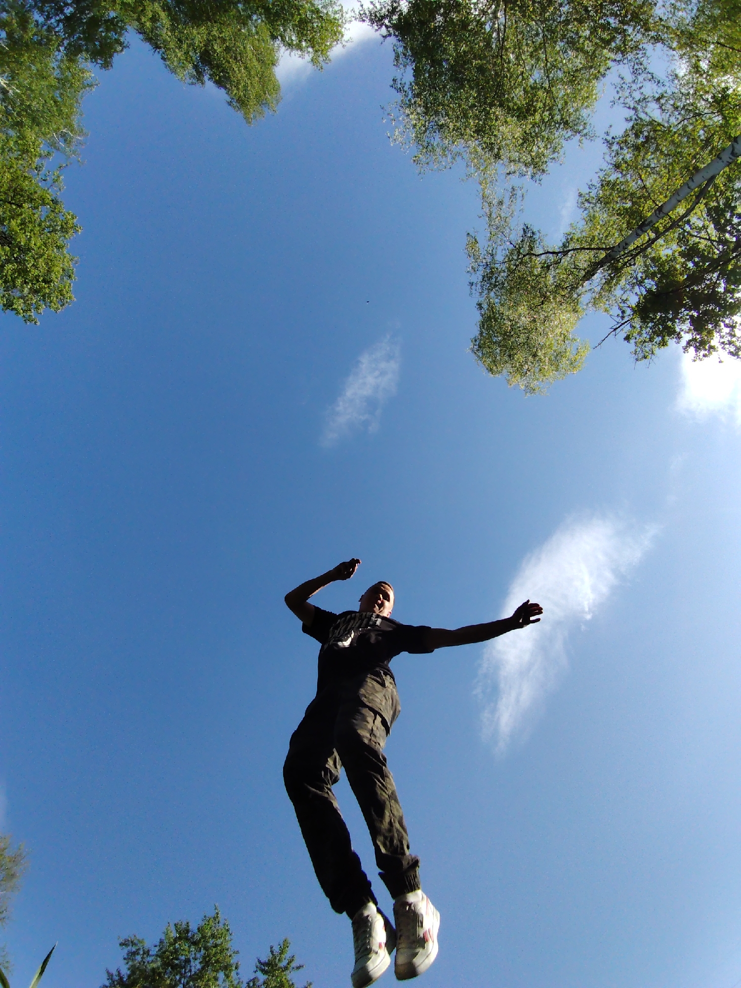 The image, taken from a low angle, shows a person in mid-air against a blue sky with scattered clouds. The person is wearing a dark t-shirt, camo-patterned pants, and white sneakers. Their arms are outstretched, suggesting they are jumping or in motion. The periphery of the image is framed by the green foliage of trees. The location is Chaikovsky, Russia.