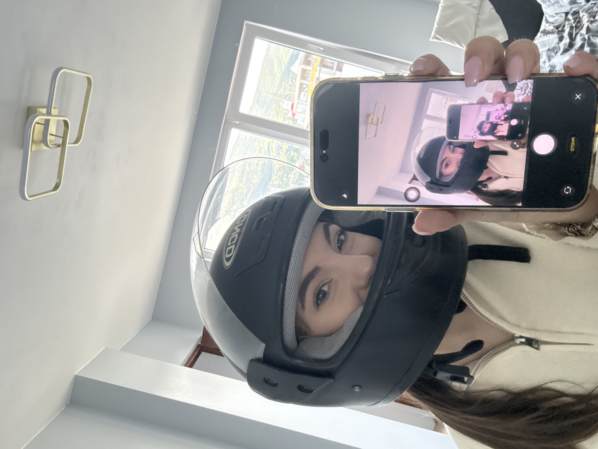 A woman wearing a black and silver motorcycle helmet is taking a photo of herself in a mirror using her smartphone. The phone screen shows a reflection of the same image, creating a visual loop. The background includes a window, a wall with a modern light fixture, and possibly an outdoor scene. The location is indoors, in a building, and the city is unknown.