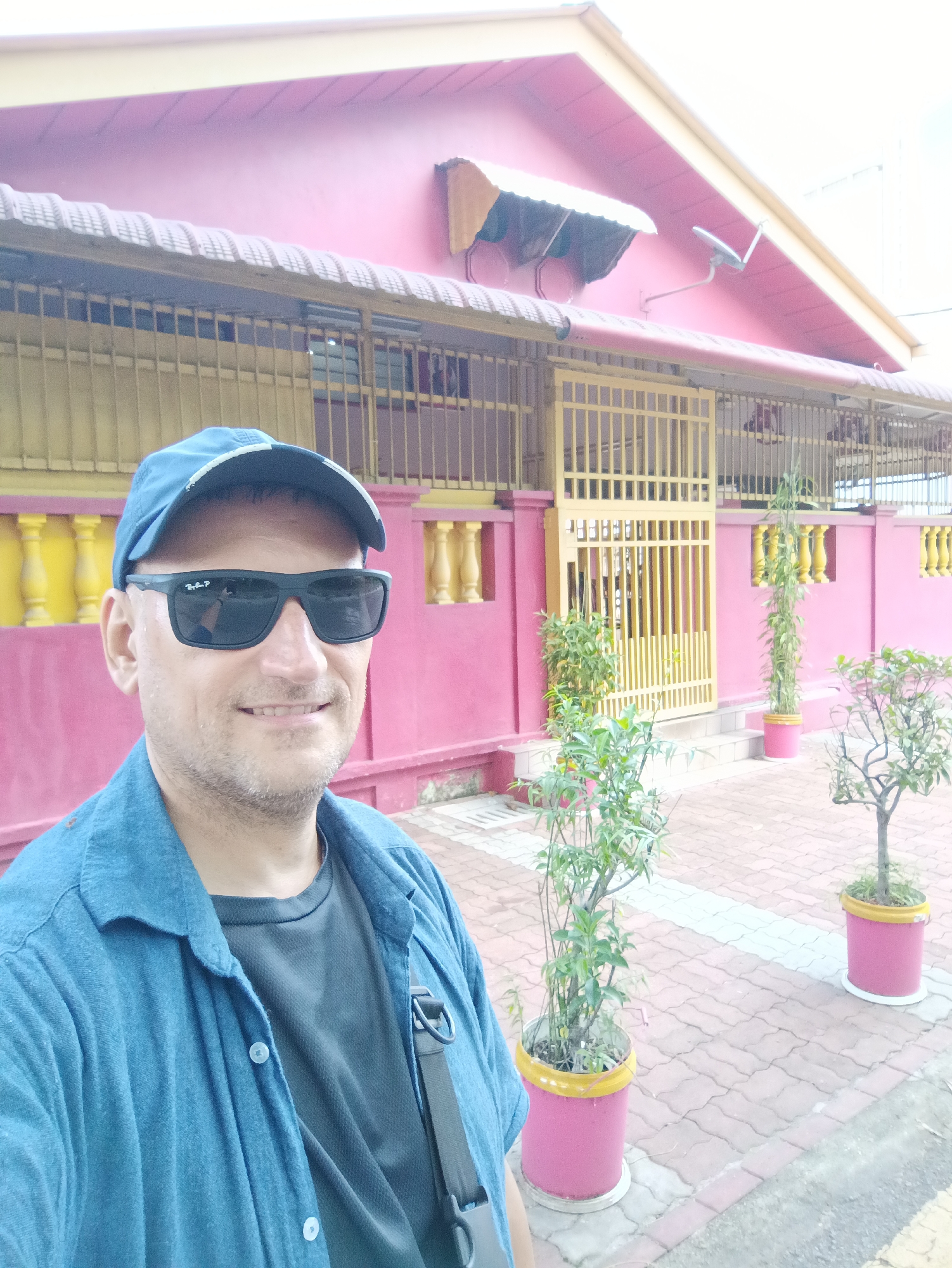 A man wearing a blue baseball cap and sunglasses takes a selfie in front of a bright pink building with a yellow fence and accents. The building appears to be a single-story structure with a sloped roof. In the foreground, there are several potted plants, including a tall, slender bamboo-like plant and a smaller, more ornamental tree. The ground is paved with interlocking bricks. The man is wearing a blue denim-like shirt over a dark t-shirt. He has a bag strap visible over his shoulder. The scene appears to be outdoors, possibly in a residential or public area.