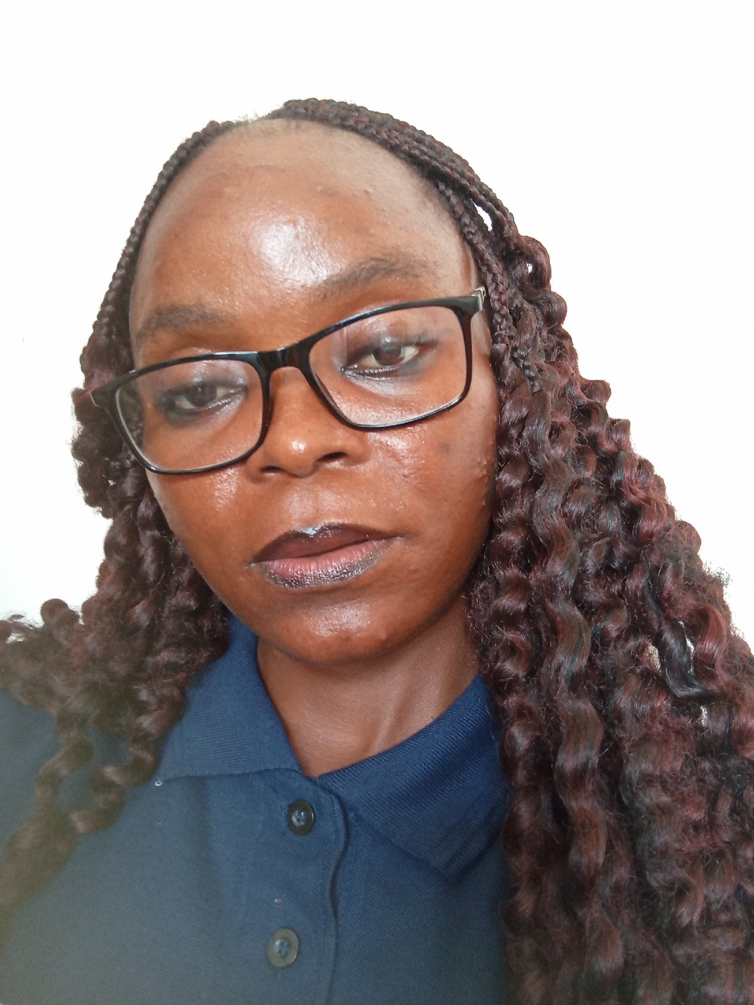 A close-up, head-and-shoulders studio portrait features a Black woman with braided hair and eyeglasses. She is looking directly at the camera, with her head tilted slightly. Her skin is dark and appears slightly oily, with some subtle texture visible. She is wearing black-rimmed glasses that frame her eyes. Her lips are painted with a dark, glossy lipstick. She is wearing a dark blue collared shirt, with two visible black buttons on the placket. The background is a plain white wall, suggesting a controlled studio or indoor setting. The lighting appears to be from the front, illuminating her face evenly. The image is an informal portrait, likely taken with a smartphone or a compact camera, given the quality and composition. There is no discernible context or specific location within the city of Jalingo, Nigeria, beyond the fact that the subject is likely a resident or visitor to the area.